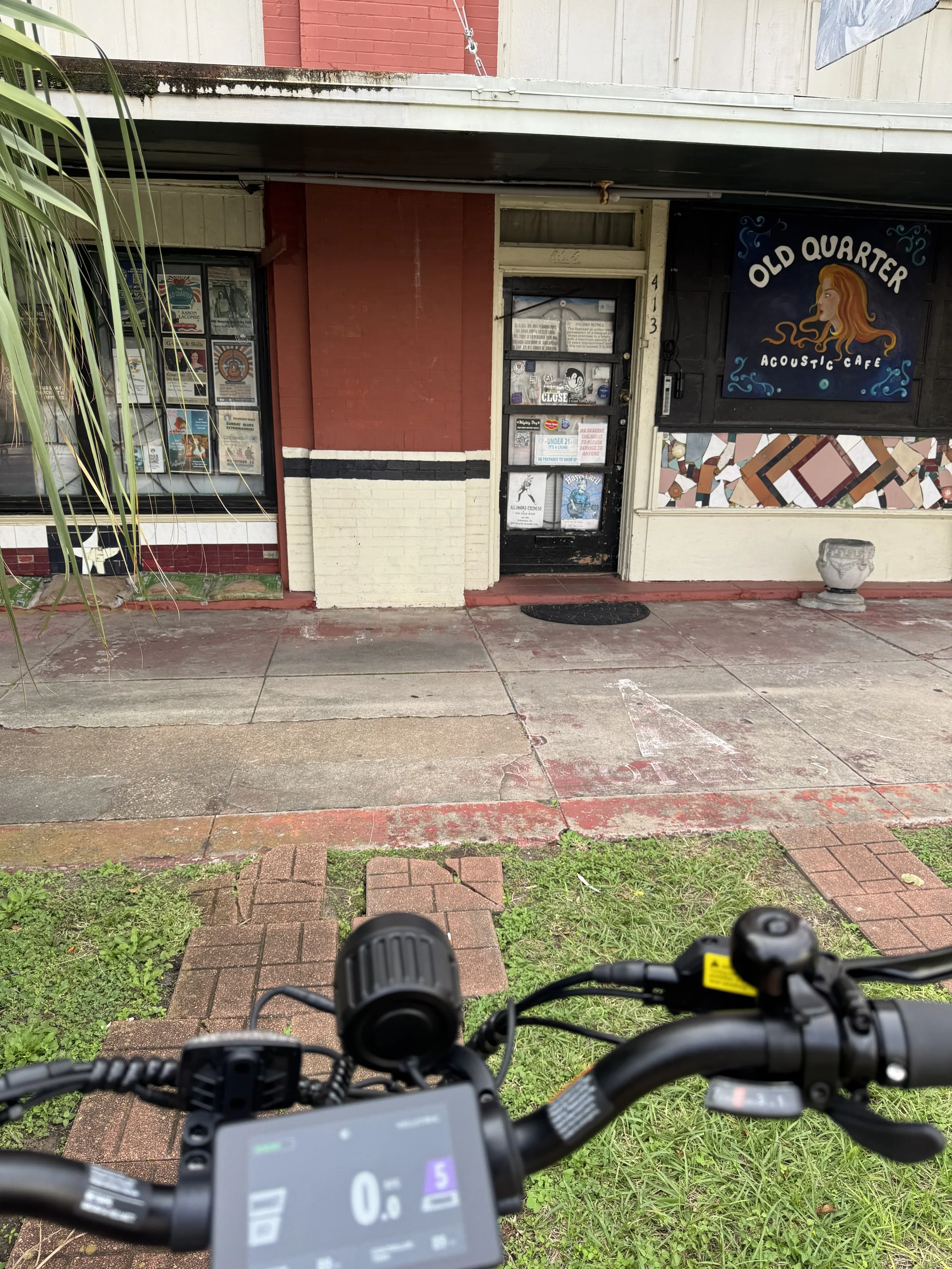 Facade of a small business called Old Quarter Acoustic Cafe with a door and large windows displaying posters. The storefront is painted in red, cream, and black, with a mosaic design on the wall to the right of the door. There is a bicycle in the foreground.