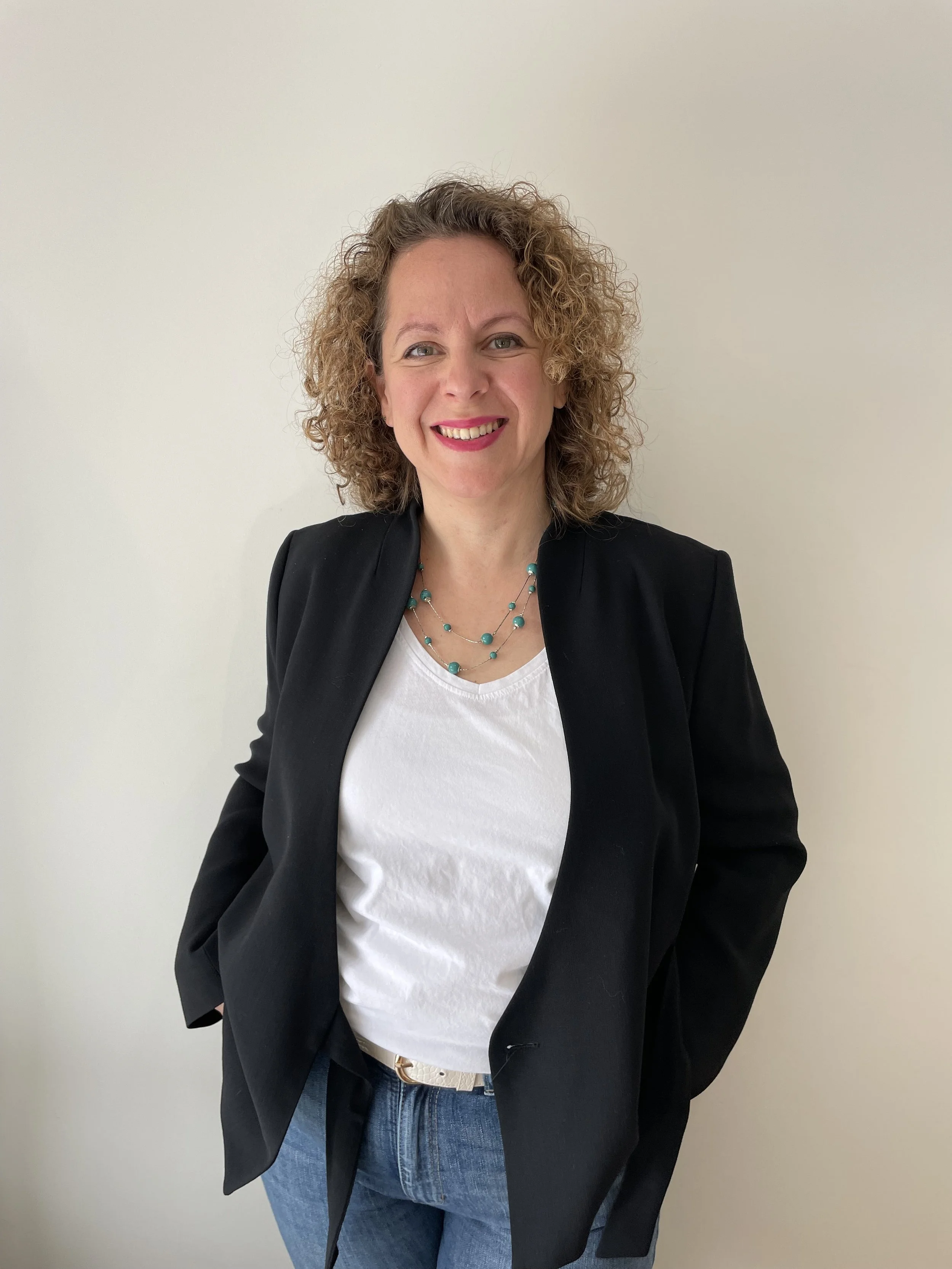 Mujer con cabello rizado, sonrisa, blazer negro, camiseta blanca, collar de piedras azules, de pie contra pared blanca.