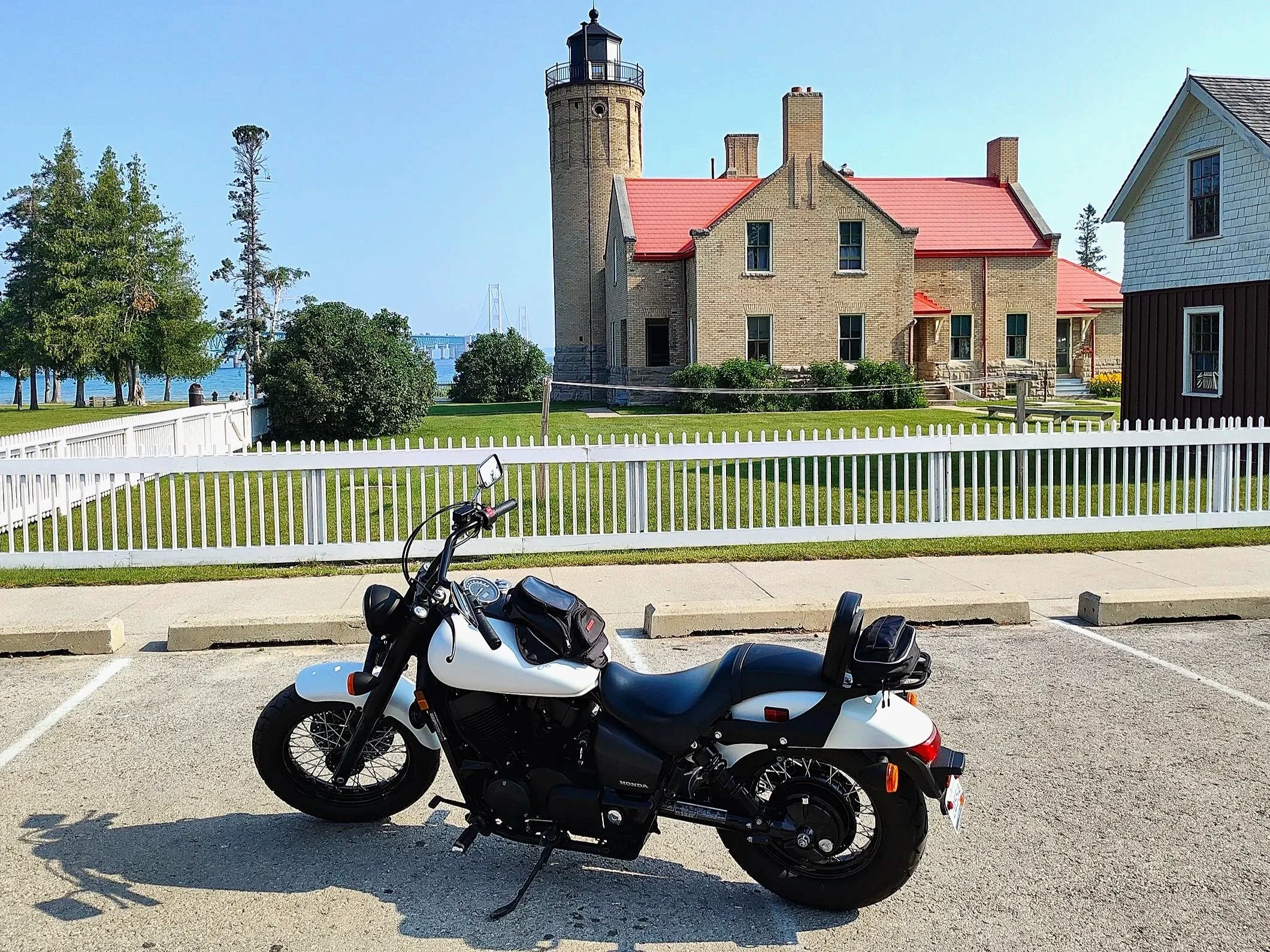 A white and black motorcycle parked in front of a white picket fence with a historic lighthouse and buildings in the background, near the water.