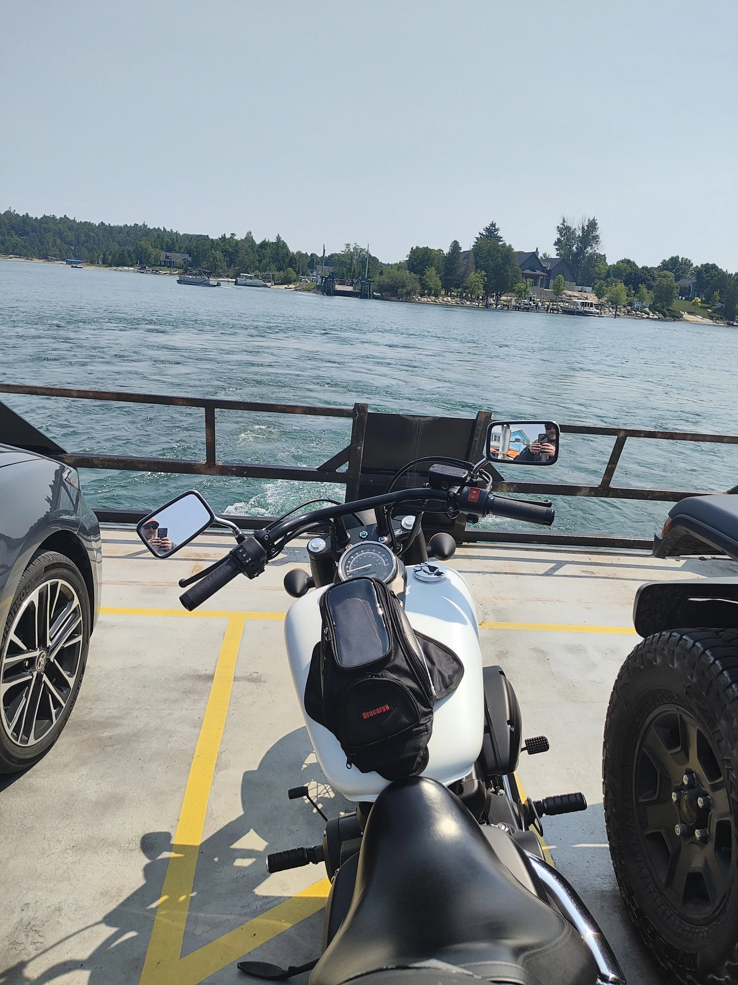 Motorcycle parked at a waterfront parking lot facing a lake with boats and houses in the distance.