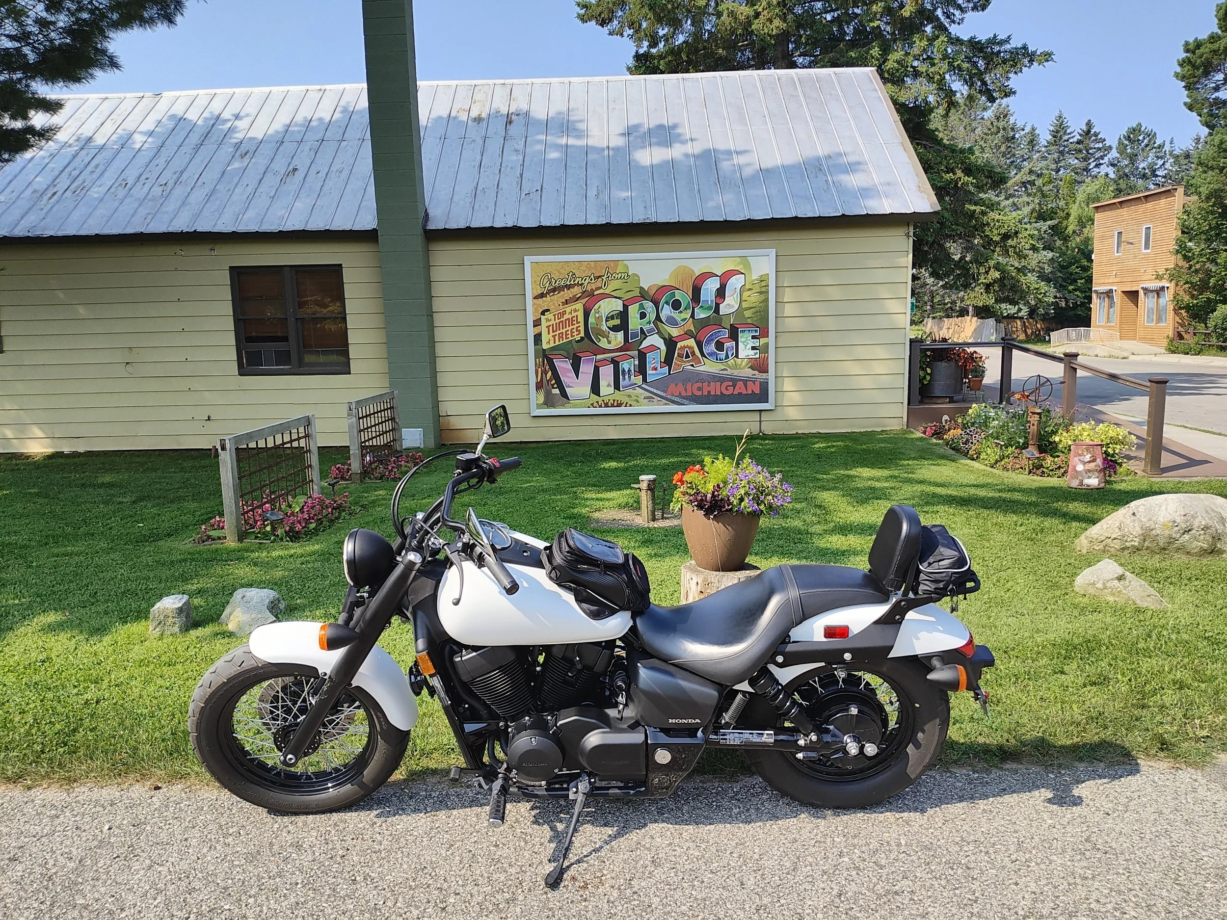 A black and white motorcycle parked on the grass in front of a yellow building with a large colorful sign that reads 'Greetings from Croft Village Michigan.' There are flowers in pots and small garden beds around the building.