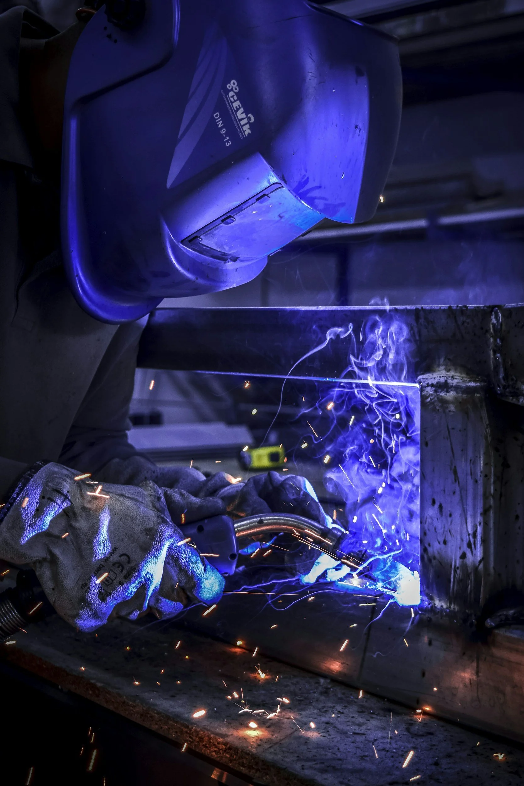A person welding metal underneath a large machine or structure, with sparks and bright blue and orange light.