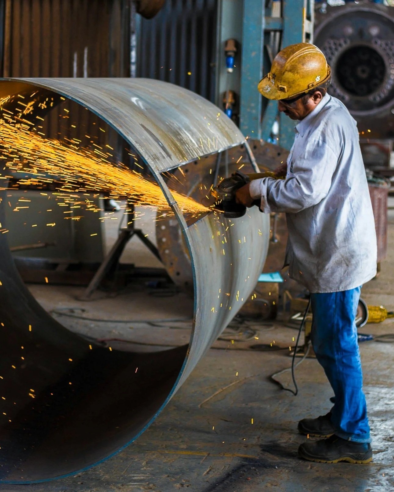 A worker wearing a yellow safety helmet and white protective coat is using an angle grinder on a large metal pipe, producing sparks in a workshop setting.