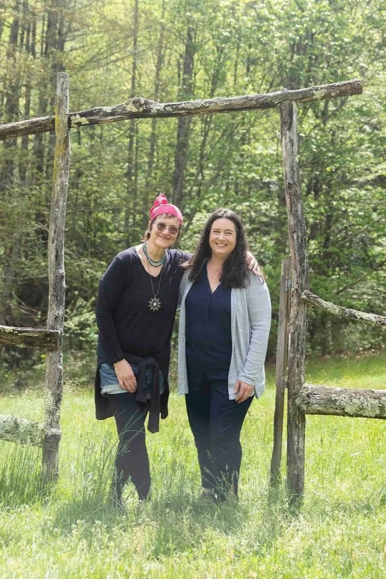 Two women standing outdoors in front of a rustic wooden frame, smiling, surrounded by trees and grass.