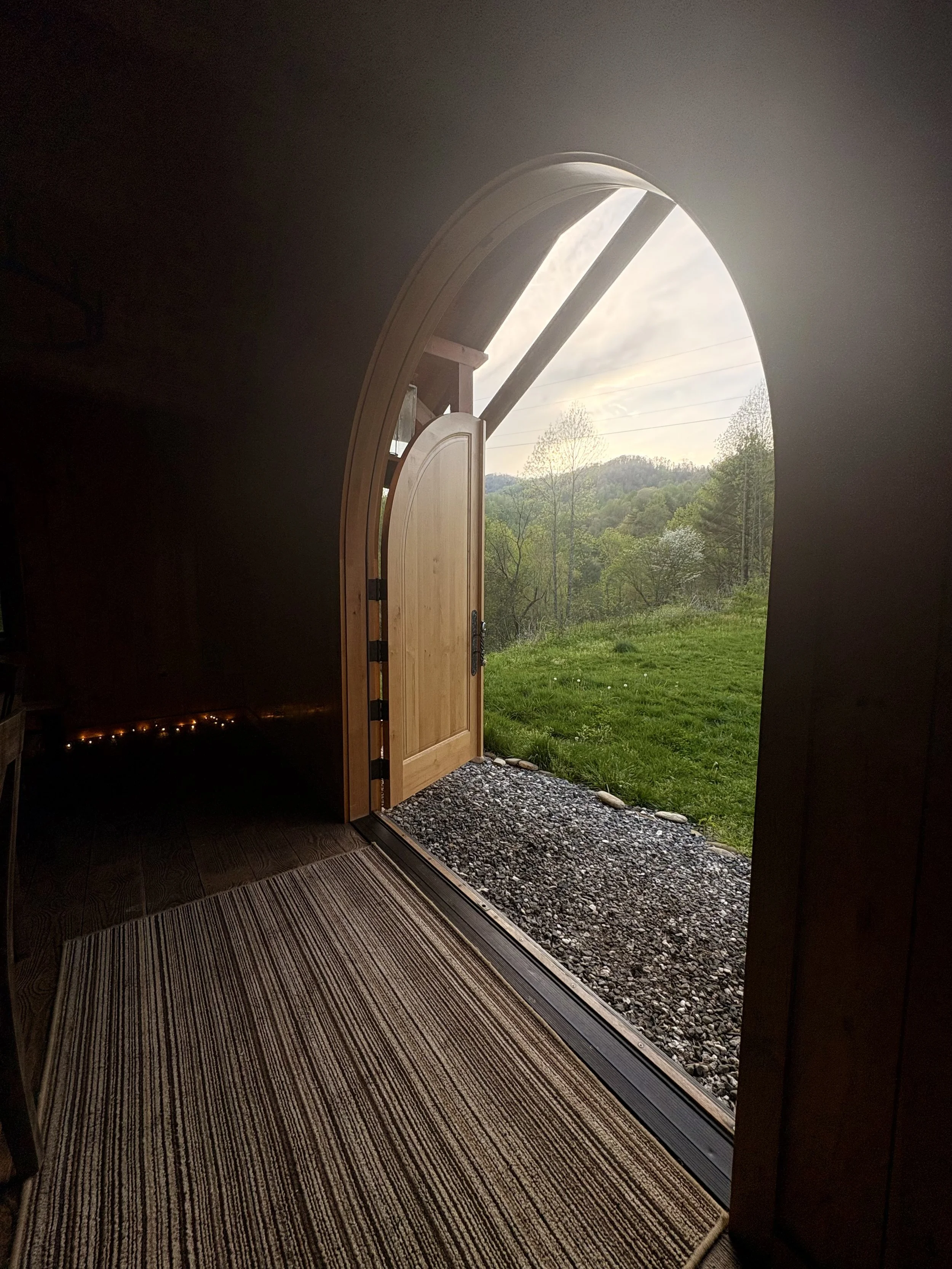 Open wooden arched door leading outside to a green grassy yard with trees and hills in the distance, under a cloudy sky.