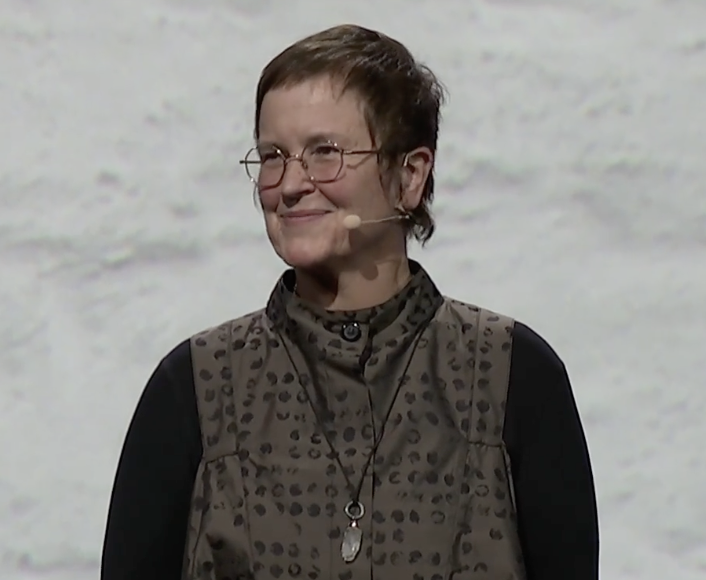 A woman with short brown hair and glasses standing on stage, wearing a black long-sleeve shirt and a patterned vest, smiling with a headset microphone.