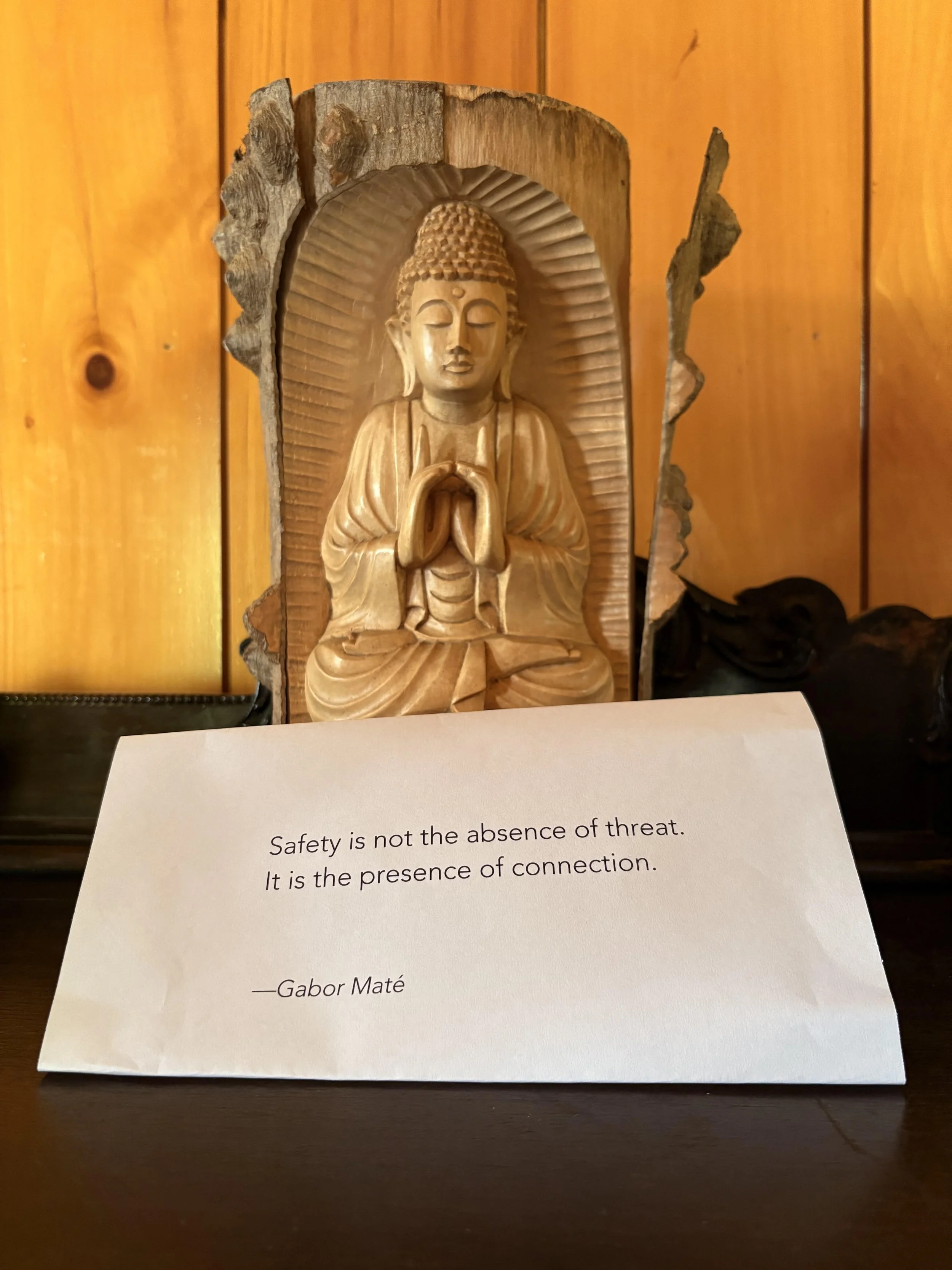 A wooden carved Buddha statue with closed eyes and hands in a prayer position, displayed against a wooden background, with a quote card in front that reads: "Safety is not the absence of threat. It is the presence of connection." - Gabor Maté.