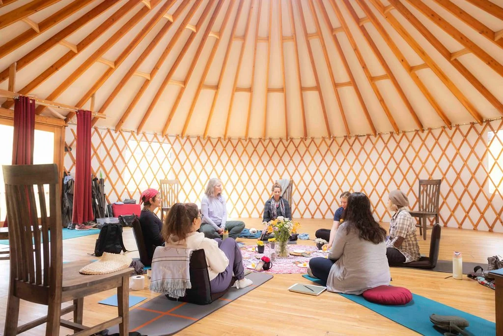 Group of people sitting in a circle inside a yurt, participating in a meditation or discussion session, with cushions, rugs, and a small table holding flowers in the center.