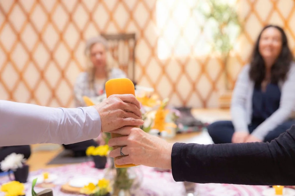 One person is handing a yellow massage roller to another person during a group activity or workshop, with two women sitting on the floor in the background.