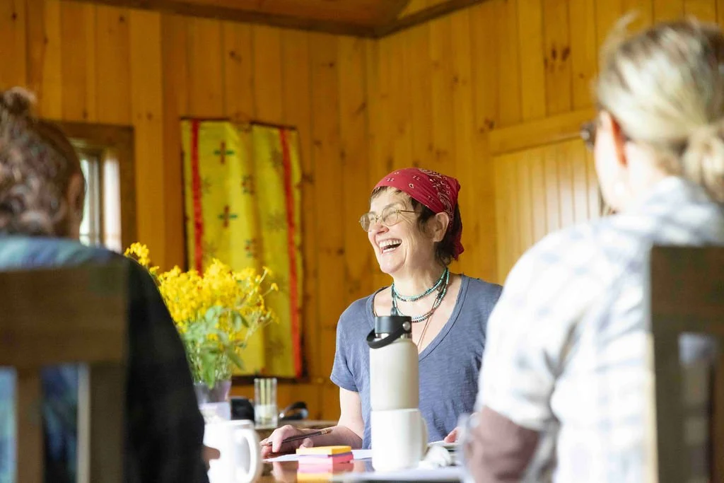 A woman with glasses and a red bandana laughing while sitting at a table with two other women in a wooden room, with flowers and a water bottle on the table.