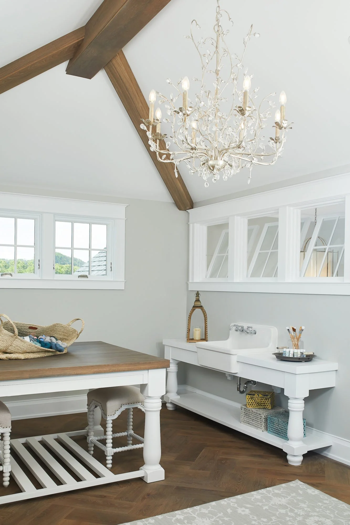 A bright white laundry or utility room with a rustic wooden-beamed ceiling, a white chandelier, a white apron sink, a window with a view of green trees, a white table with a woven basket, and colorful storage baskets underneath the sink.