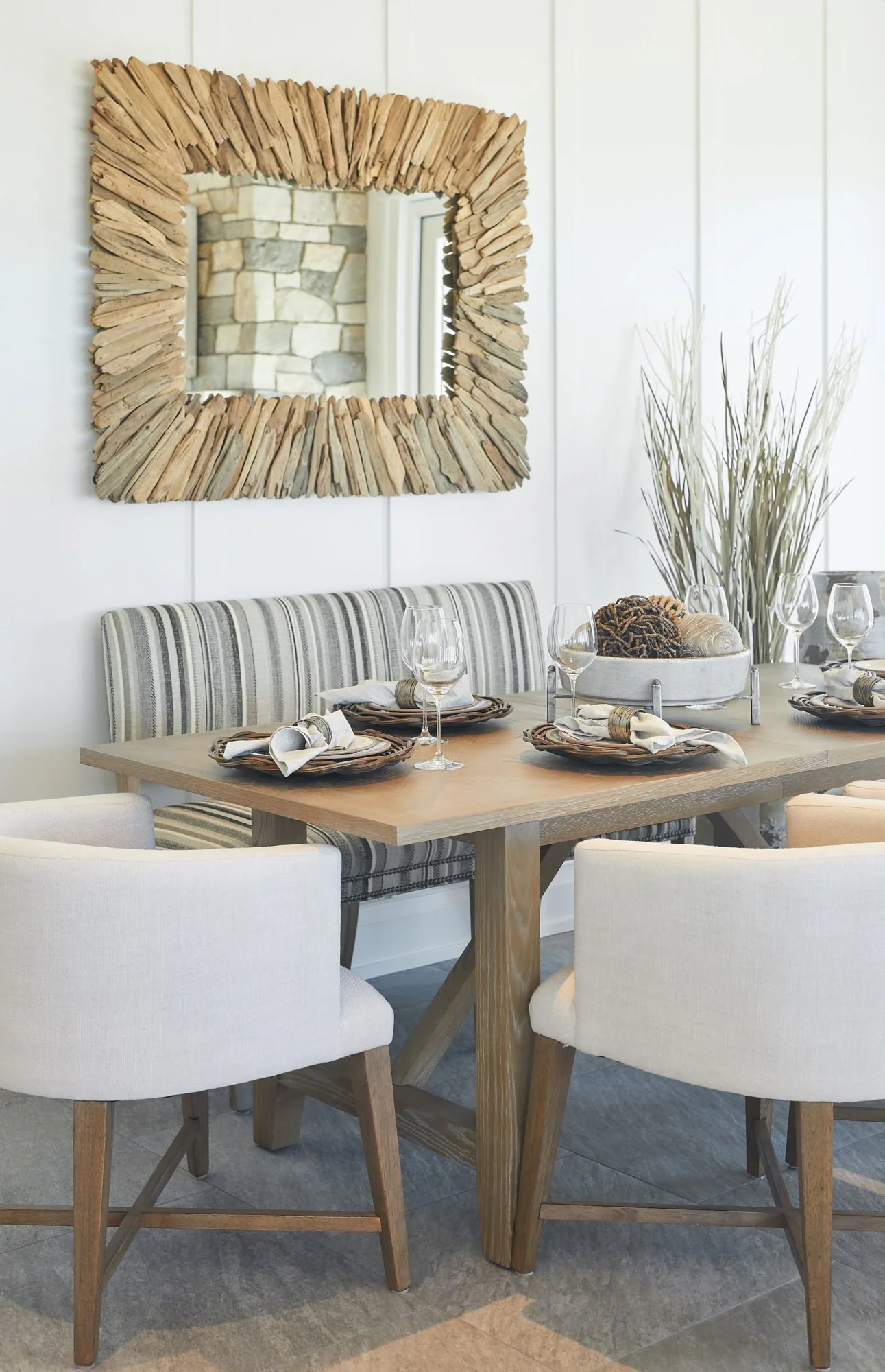 A dining room with a wooden table set with four place settings, including plates, napkins, and wine glasses. There are two white cushioned chairs and a bench with striped upholstery. A large round tray on the table holds decorative items. Behind the 