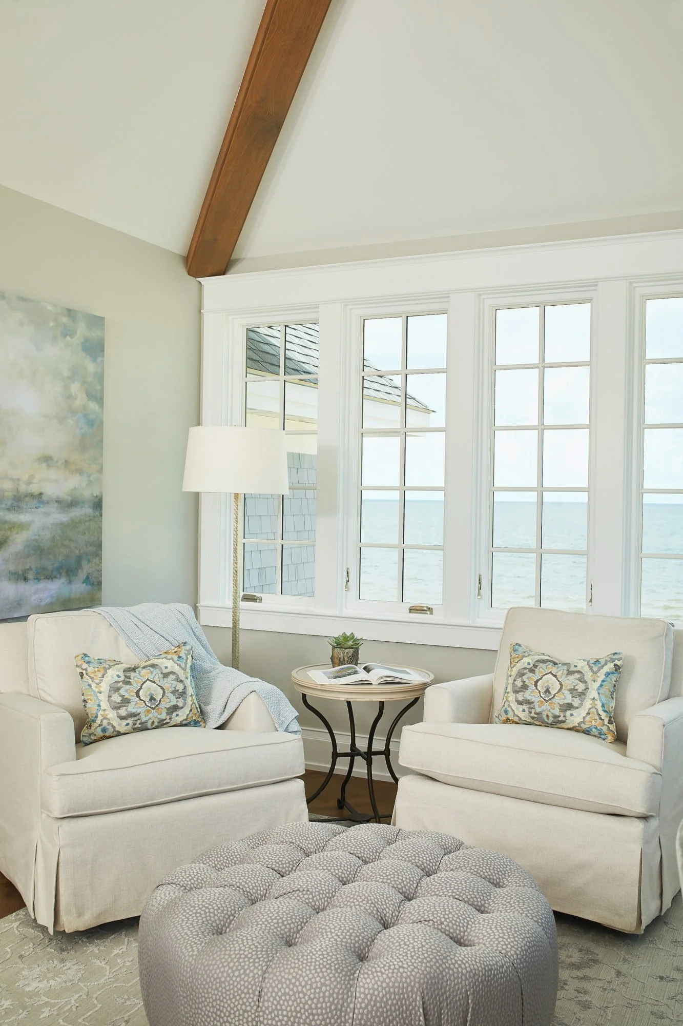Cozy living room with two white armchairs, a round tufted ottoman, a small side table with a book and a potted plant, large windows with ocean view, and a floor lamp.