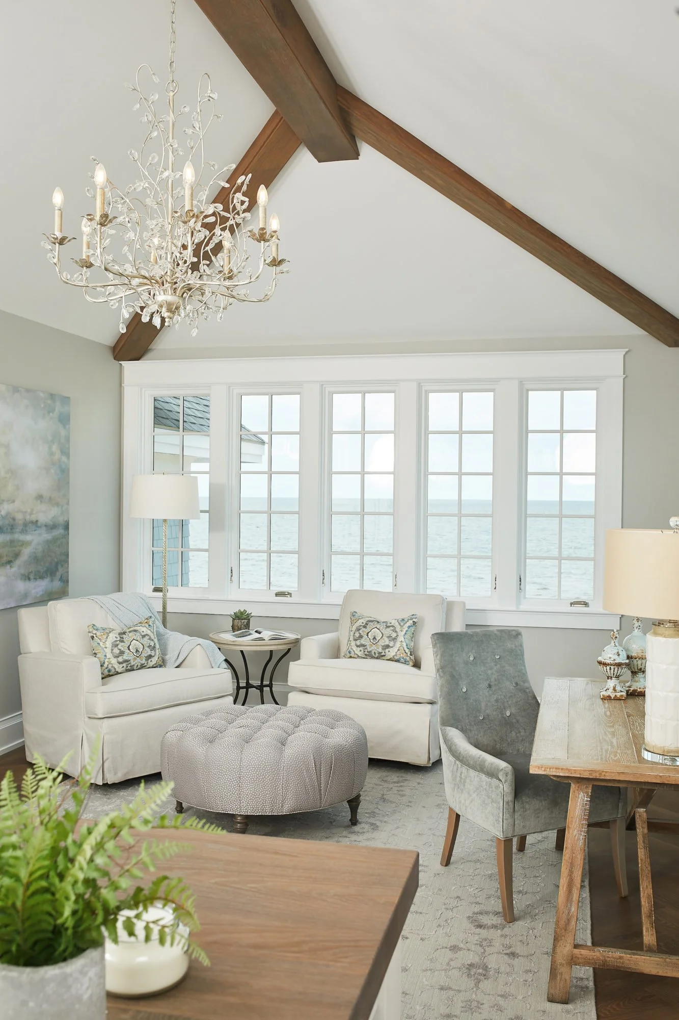 Coastal living room with white couches, gray armchair, tufted ottoman, wooden dining table, fireplace, large window view of the ocean, chandelier, and decorative pillows.