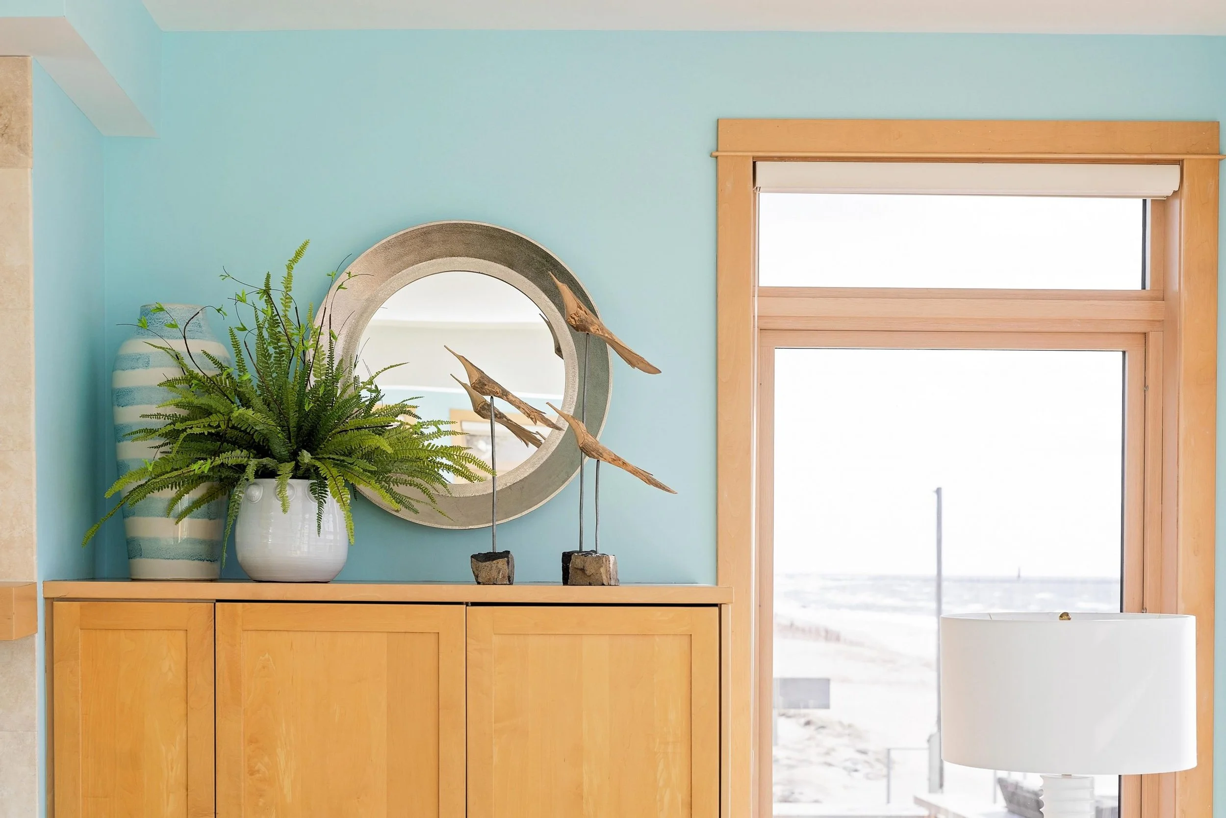 Decorative interior with wooden cabinet, mirror, large plant, striped vase, wooden birds sculpture, window with a view of the beach and white lamp.