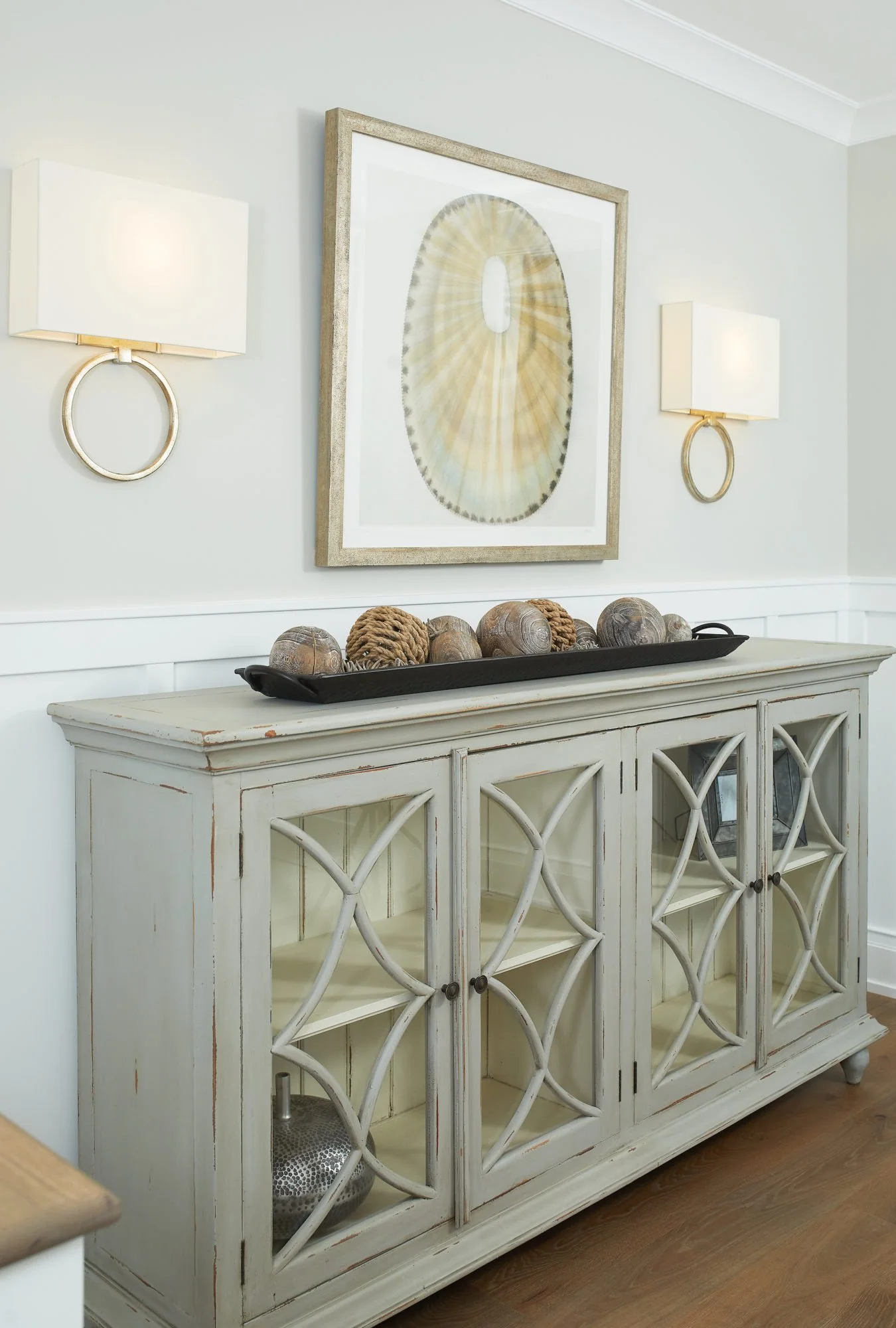 A white distressed sideboard with decorative white lattice doors, topped with a black tray holding seashells and coral arrangements. Above, a framed artwork of a sand dollar and two wall-mounted gold and white lamps are displayed on a light gray wall