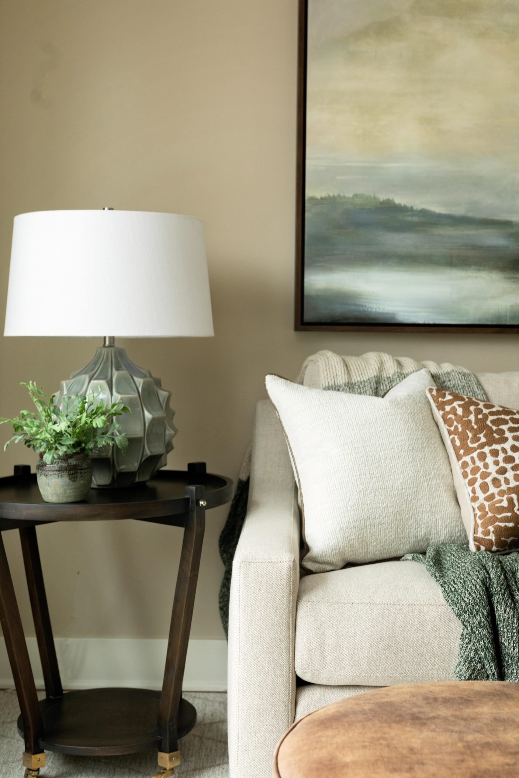 Living room corner with a beige sofa, decorative pillows, a dark wooden side table, a white lamp, green plant, and a landscape painting on the wall.