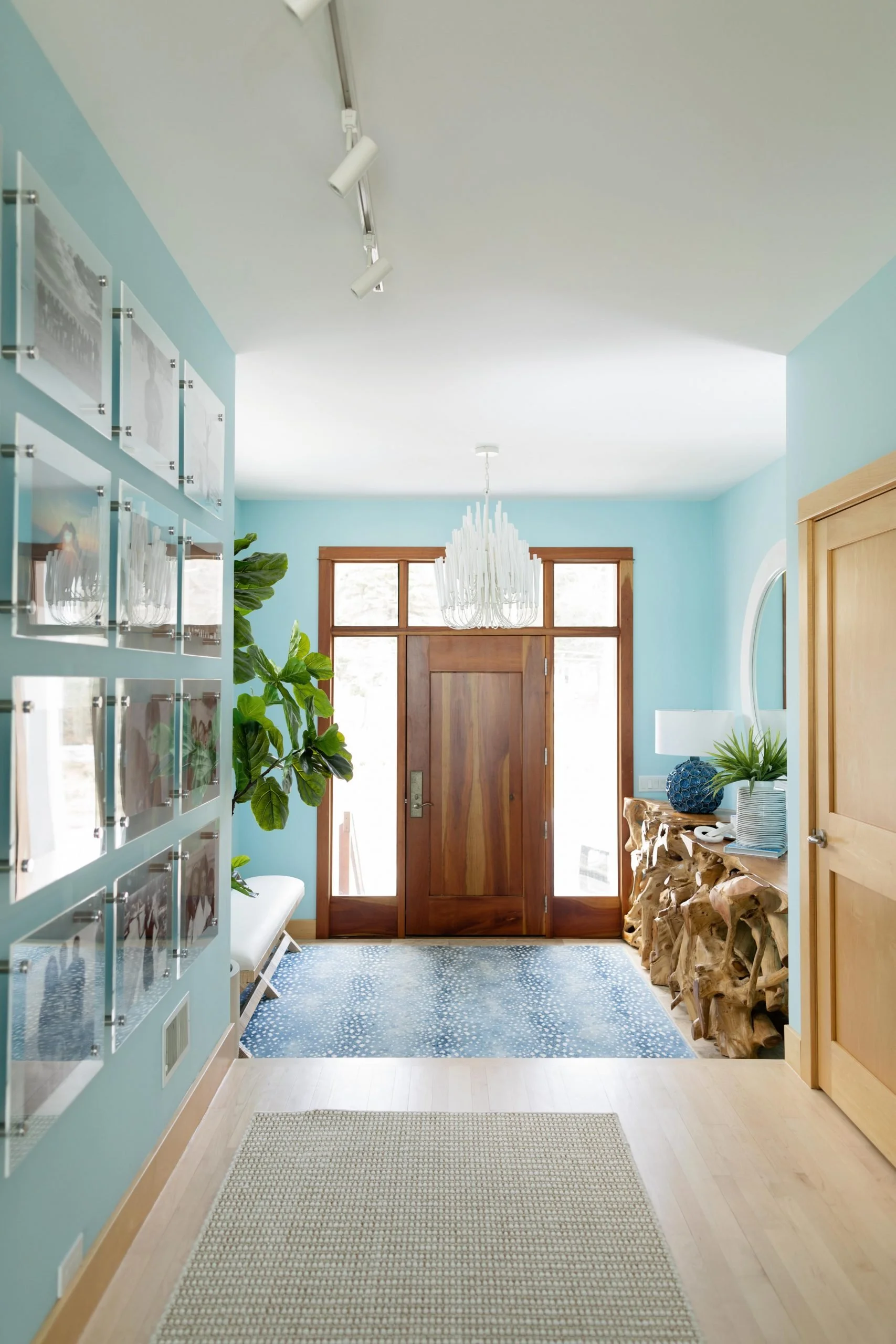 Entryway with wooden front door, light blue walls, potted plant, chandelier, decorative wooden side table with blue vase and plants, and a wall with photo frames.