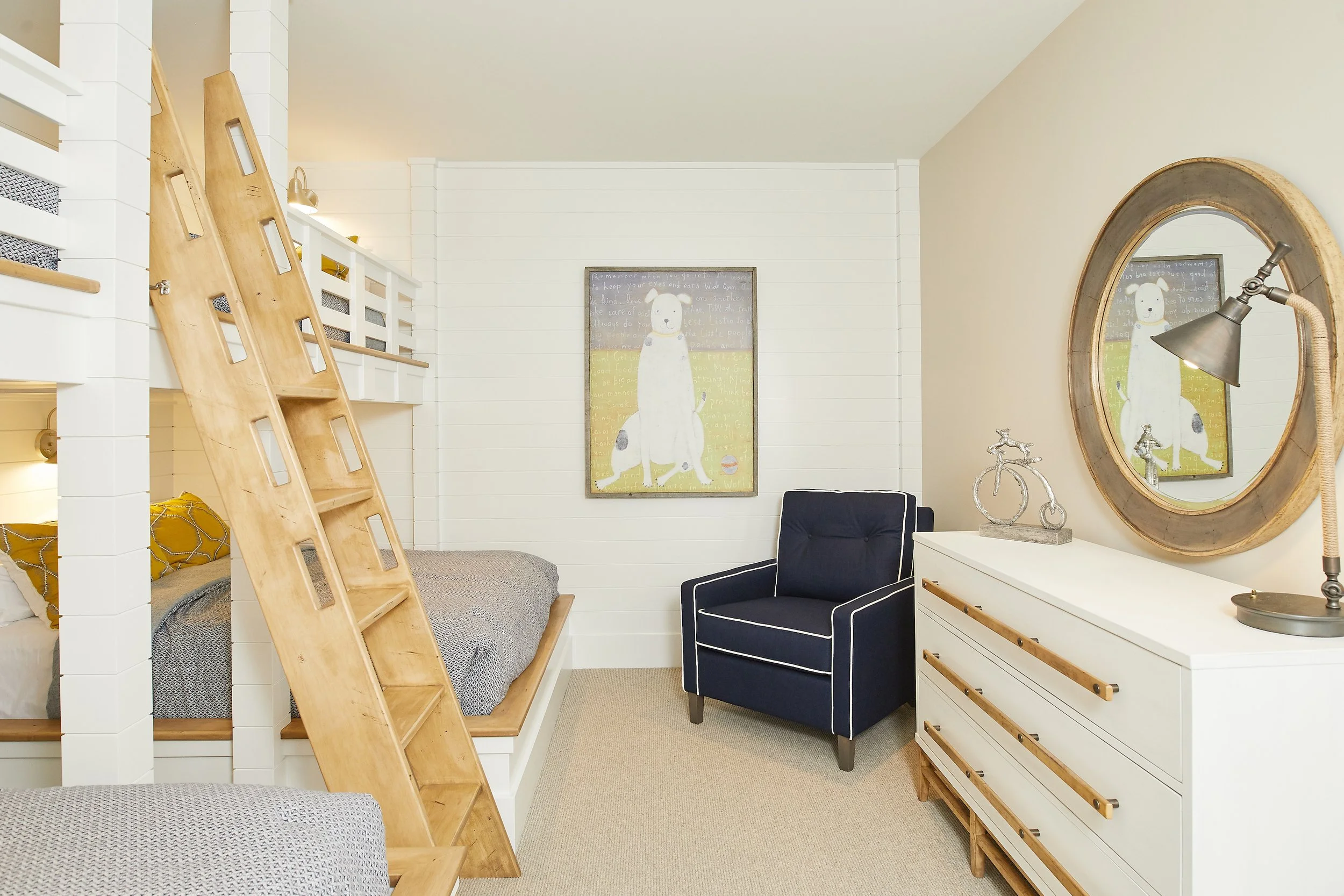 A cozy bedroom with a bunk bed, a ladder, a navy blue armchair, a white dresser with a lamp and decorative bicycle, a round mirror, and wall art of a dog.