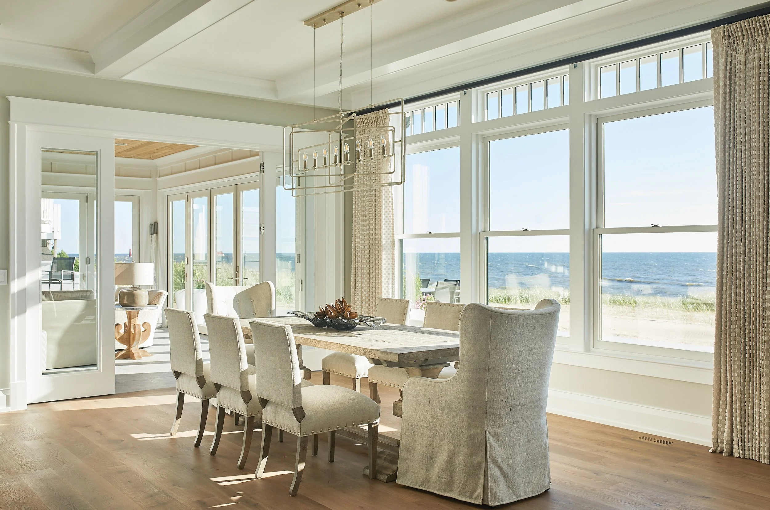 Dining room with large windows overlooking the beach and ocean, a wooden table with six upholstered chairs, and a modern chandelier.