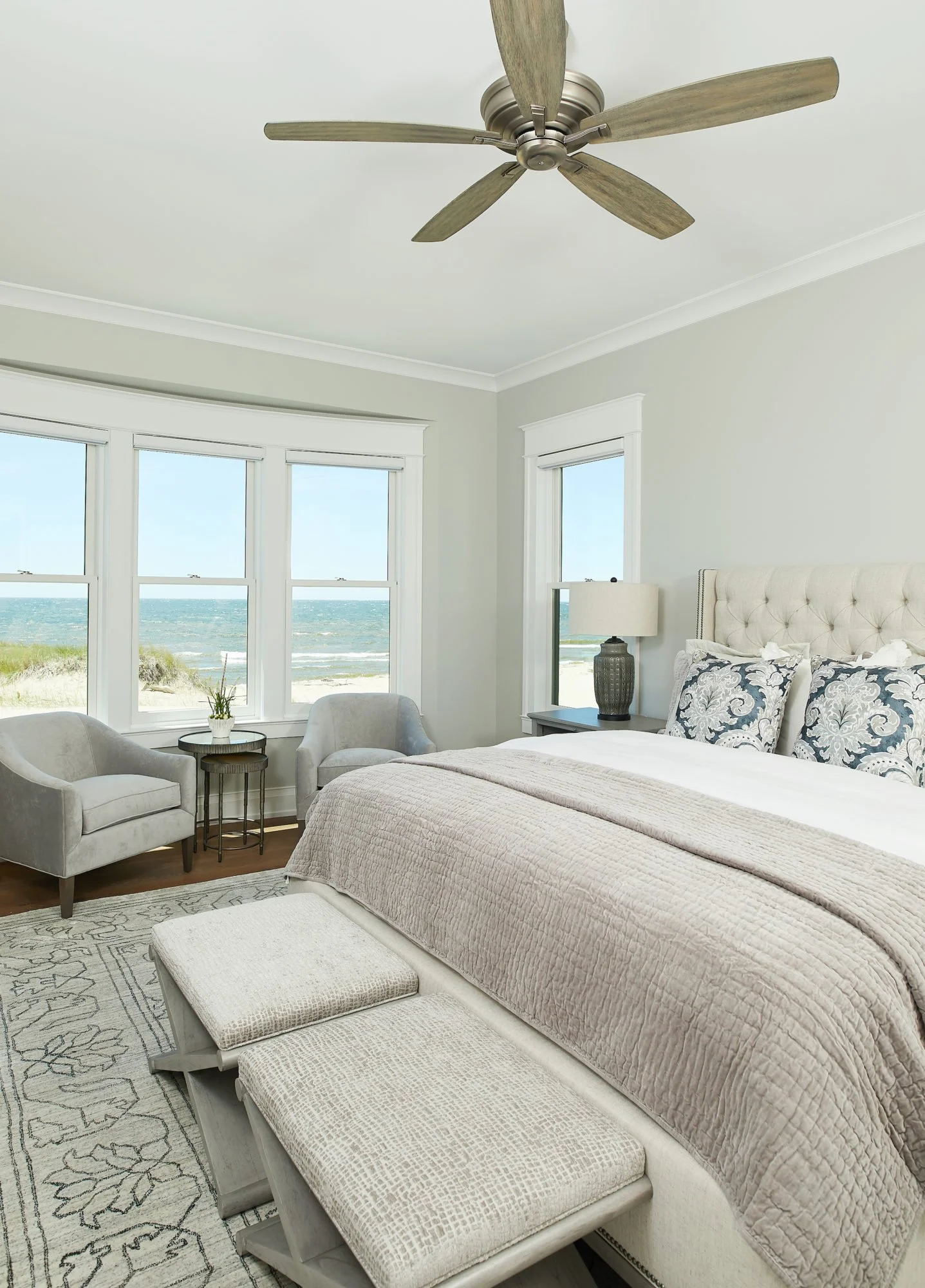 A bedroom with large windows overlooking a beach and ocean, featuring a beige bed with patterned pillows, a nightstand with a dark lamp, two gray armchairs with a small round table, and a ceiling fan.