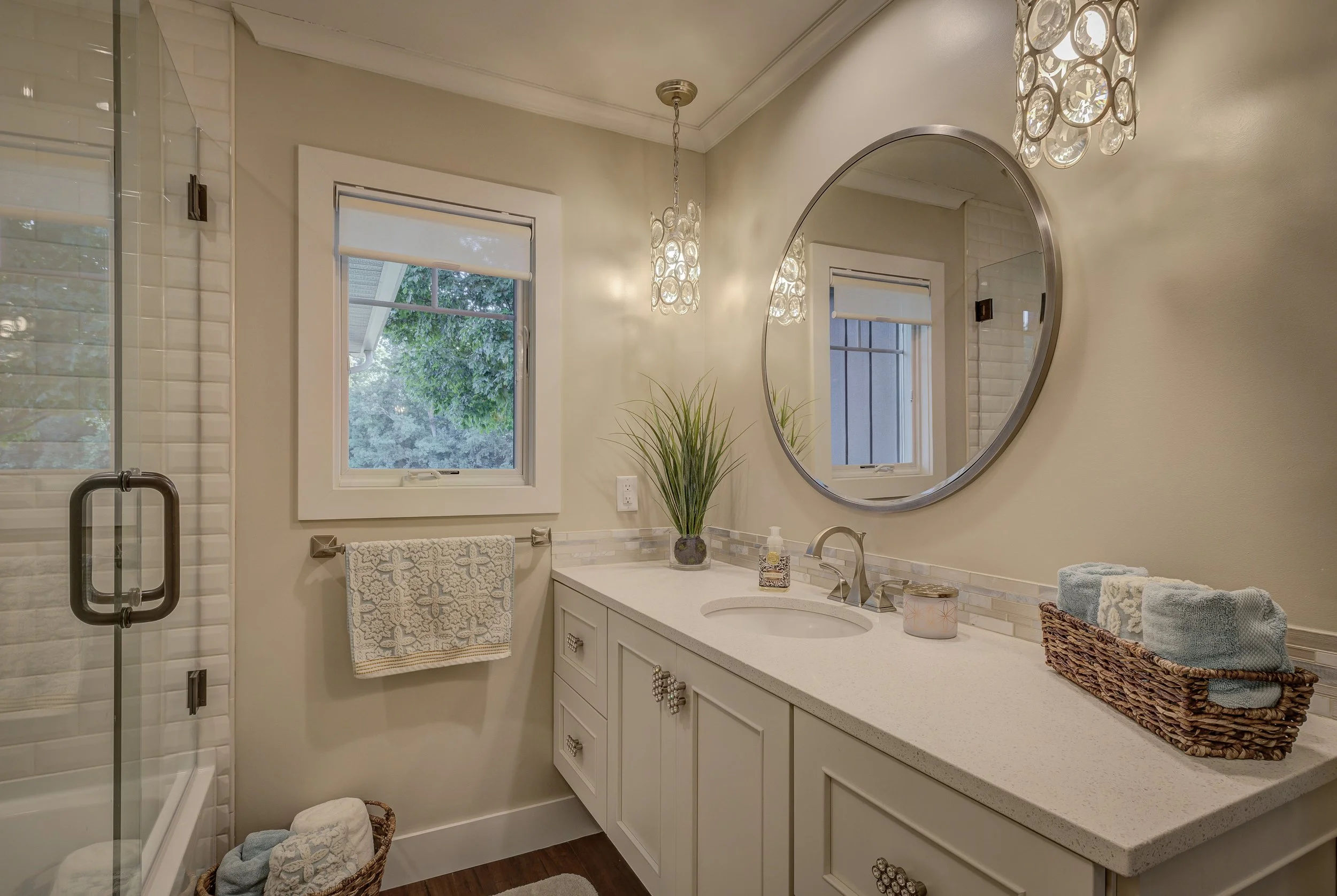 Bathroom with a white vanity, oval mirror, window showing greenery outside, towels, glass shower door, and decorative lighting fixtures.