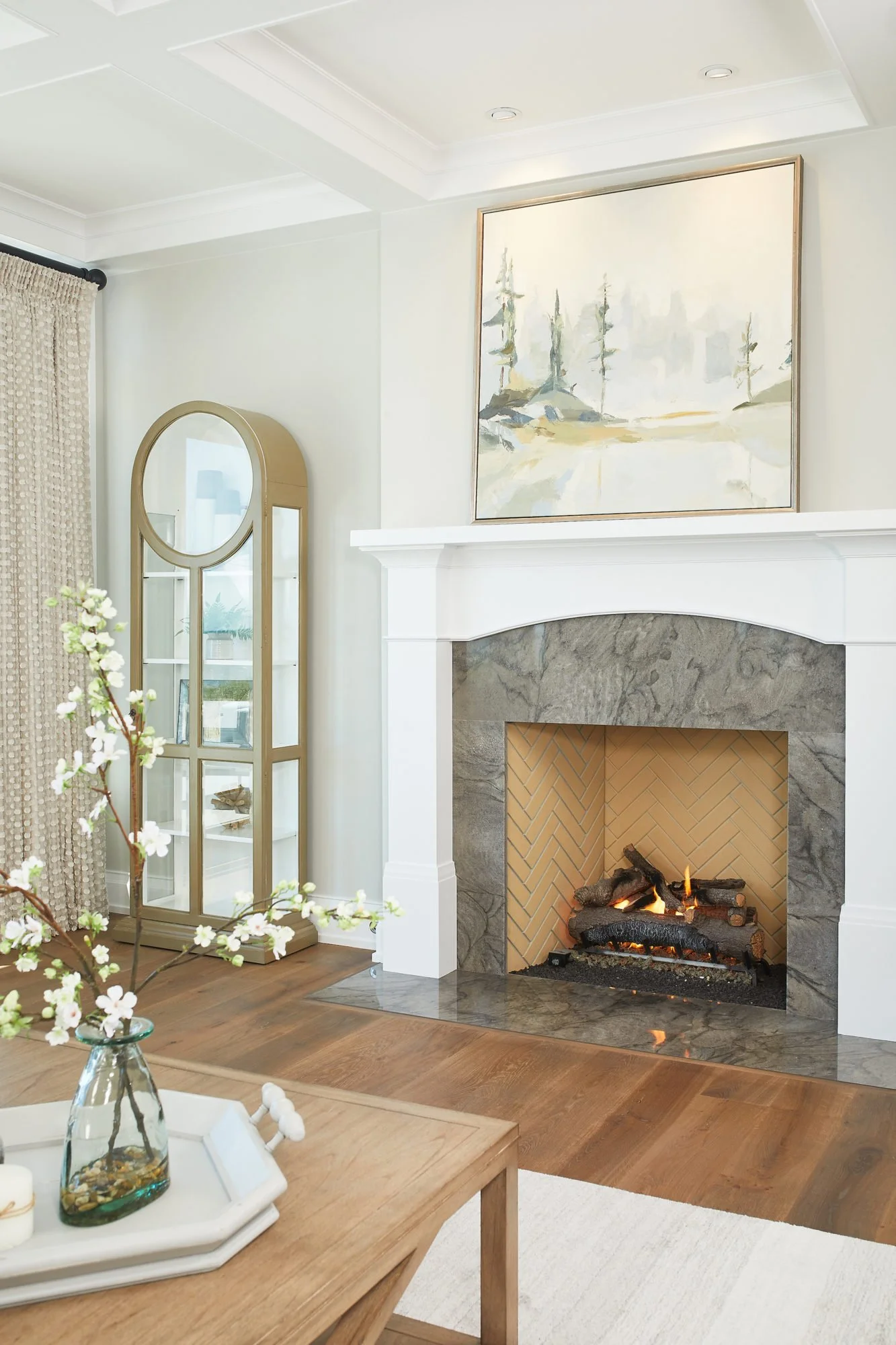 Living room with a fireplace, painting above the mantel, and a glass cabinet to the left. A table with a vase of flowers is in the foreground.