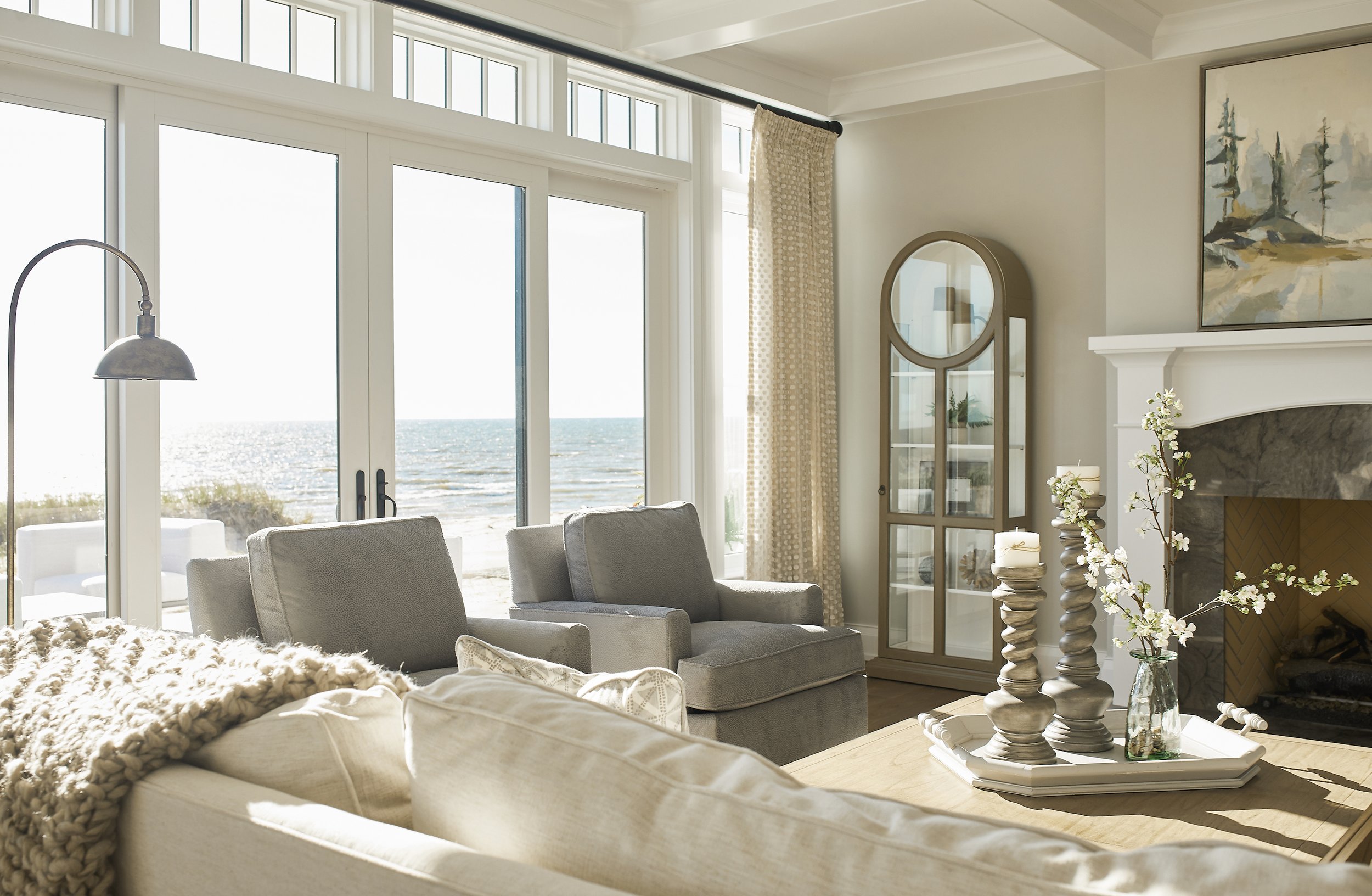 Bright living room with large glass doors showing a view of the ocean, beige and gray furniture, a fireplace with candles and decorative candles on carved holders, a glass cabinet with plants, and artwork on the wall.