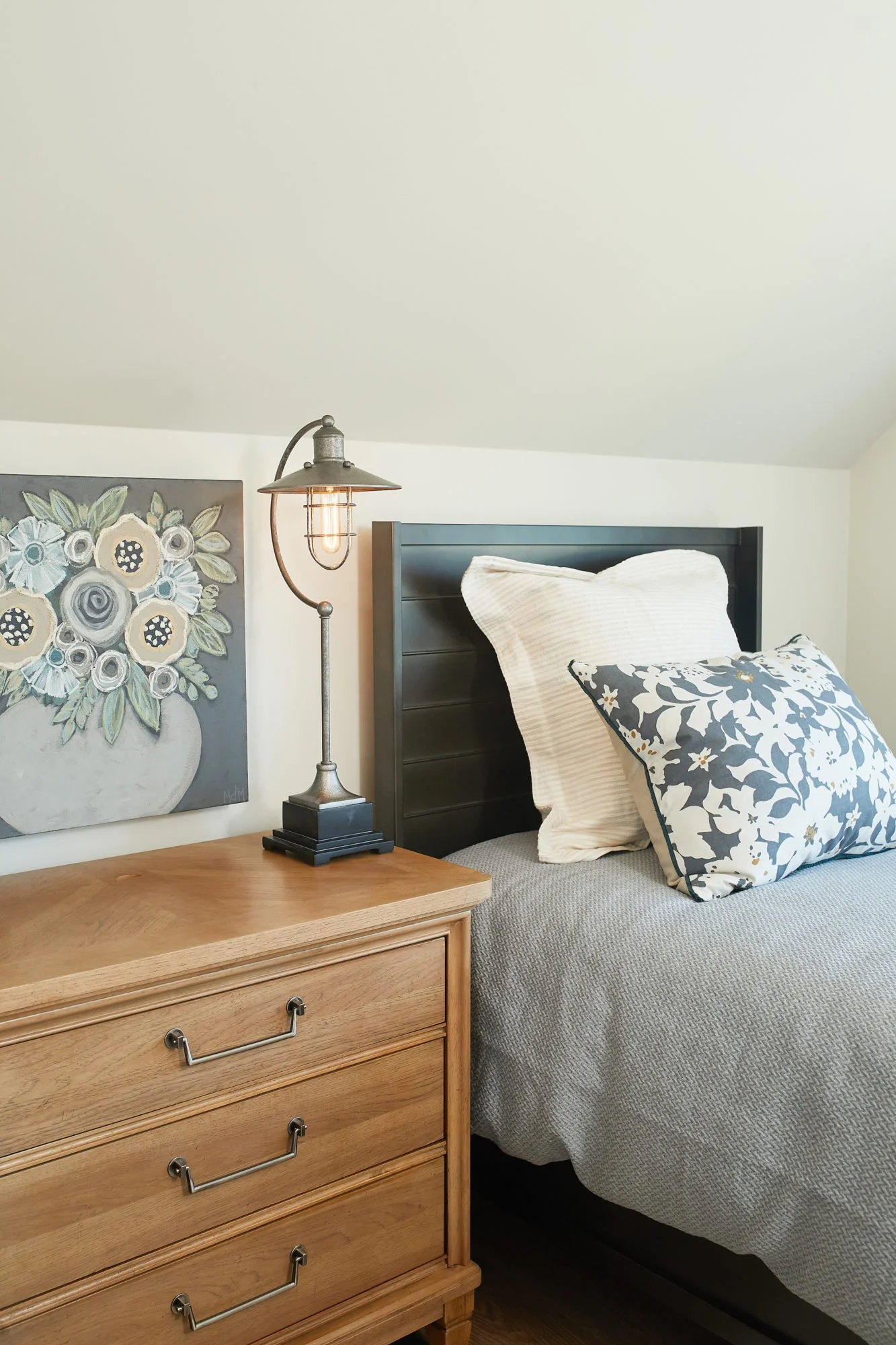 A bedroom corner featuring a wooden dresser with brass handles, a black bedside lamp with an exposed bulb, a large floral painting, and a bed with black headboard, white and patterned pillows, and a gray bedspread.