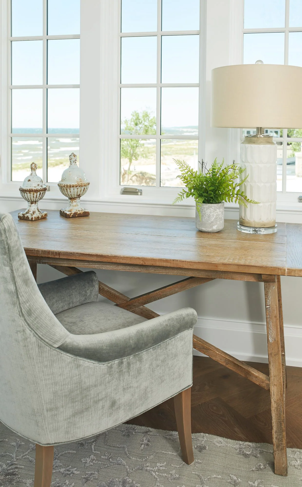 A cozy desk corner with a wooden desk, a beige velvet armchair, a classic white lamp, a small potted green fern, and two vintage decorative jars, all near large windows with a scenic outdoor view.
