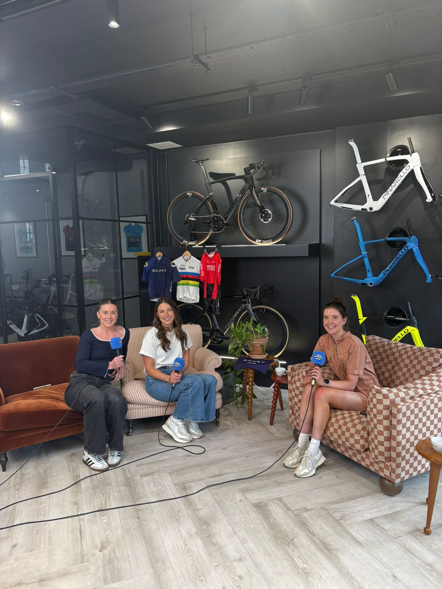 Three women sitting on vintage sofas inside a room decorated with mounted bicycles and cycling jerseys, holding microphones, with a potted plant on a small table between them.