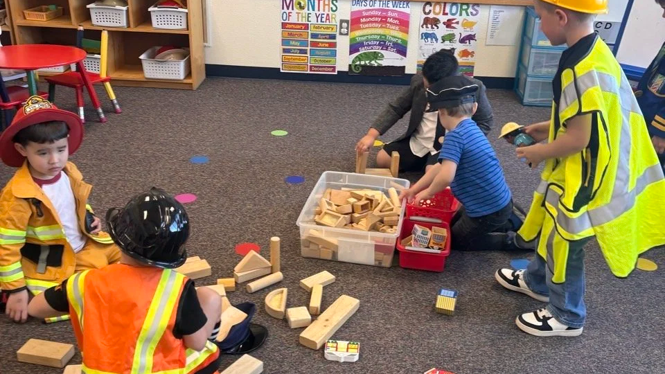 Children dressed as firefighters playing with wooden building blocks on a classroom carpet.
