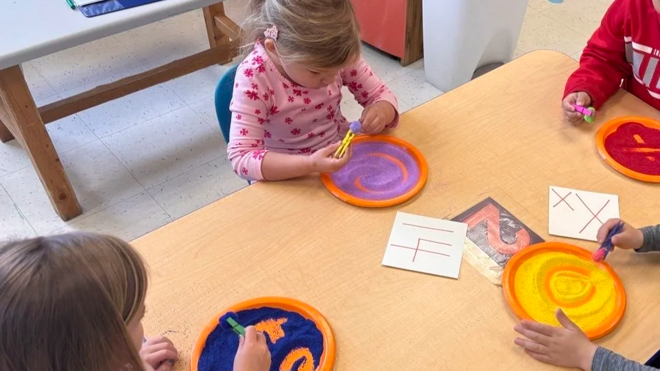 Four children sitting around a table playing with sand art kits and drawing on paper with markers in a classroom.