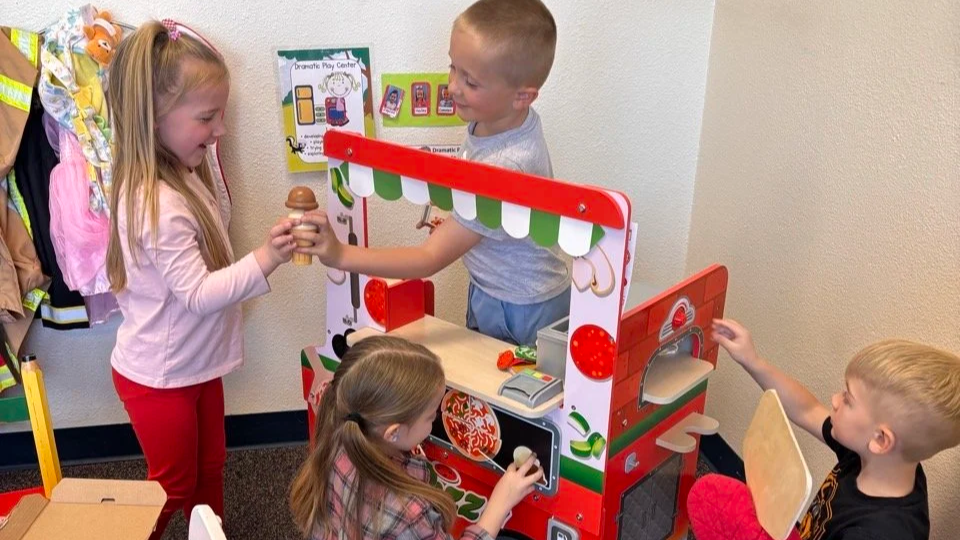 Three children playing a pretend food vendor game at an indoor play area, with a girl passing an ice cream cone to a boy, while a girl and boy sit nearby.