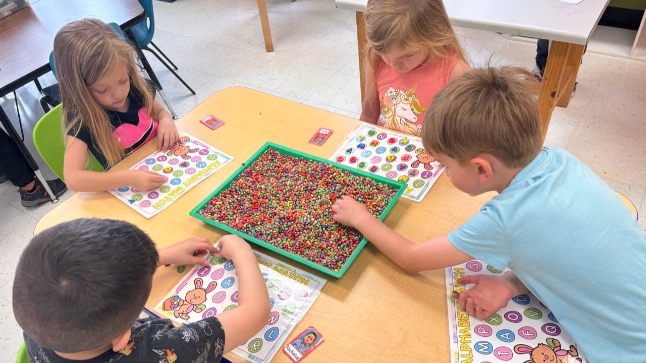 Four children playing a game with colorful small beads at a table, with alphabet-themed activity sheets and trading cards.