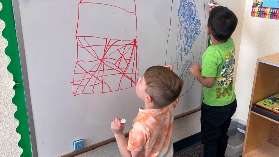 Two young boys drawing with markers on a whiteboard in a classroom.