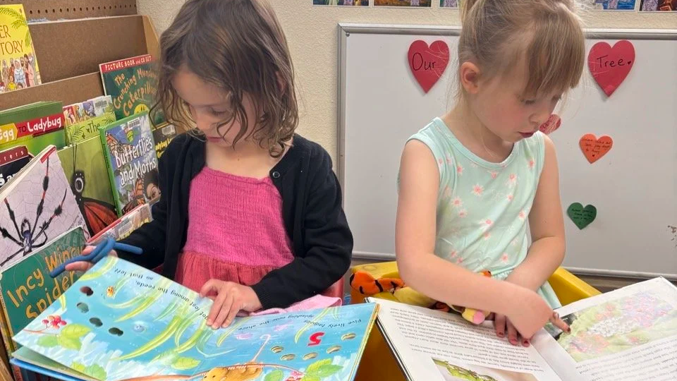 Two young girls looking at picture books in a classroom setting, with a whiteboard and colorful hearts on the wall in the background.