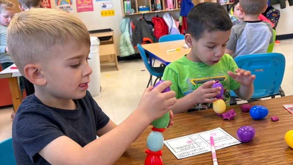 Two young boys playing with colorful plastic eggs and toys at a table in a classroom setting.