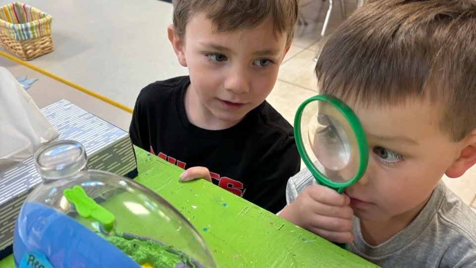 Two young boys looking intently at a plastic terrarium, one boy using a magnifying glass to observe it closely.