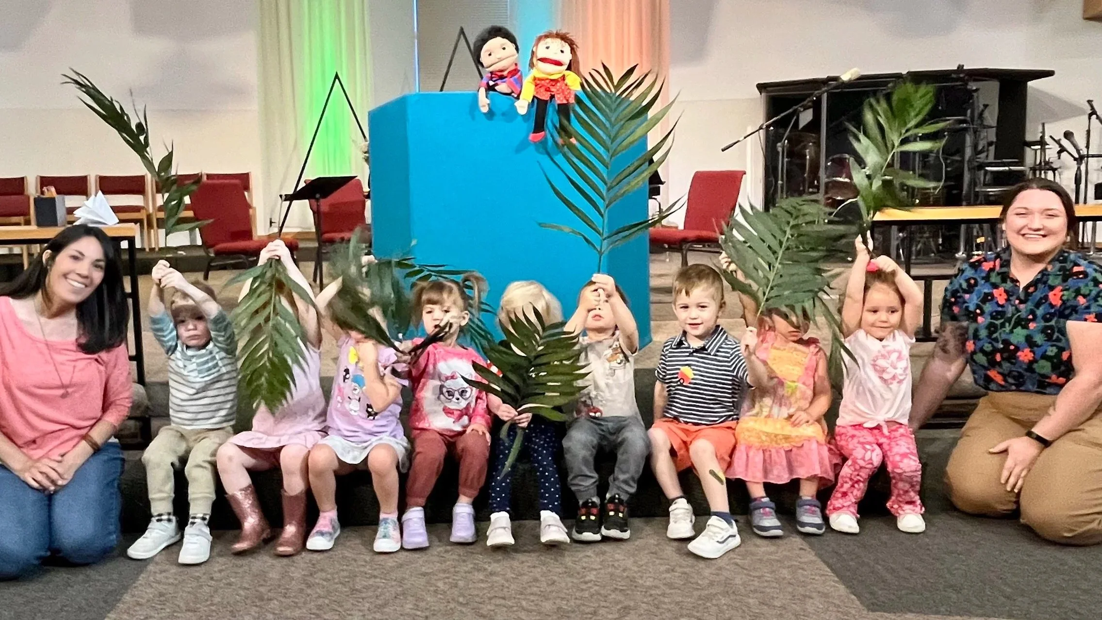 Children sitting on a stage holding large palm leaves with two women, one on each side, smiling. Puppets are placed on a blue rectangular block in the background.