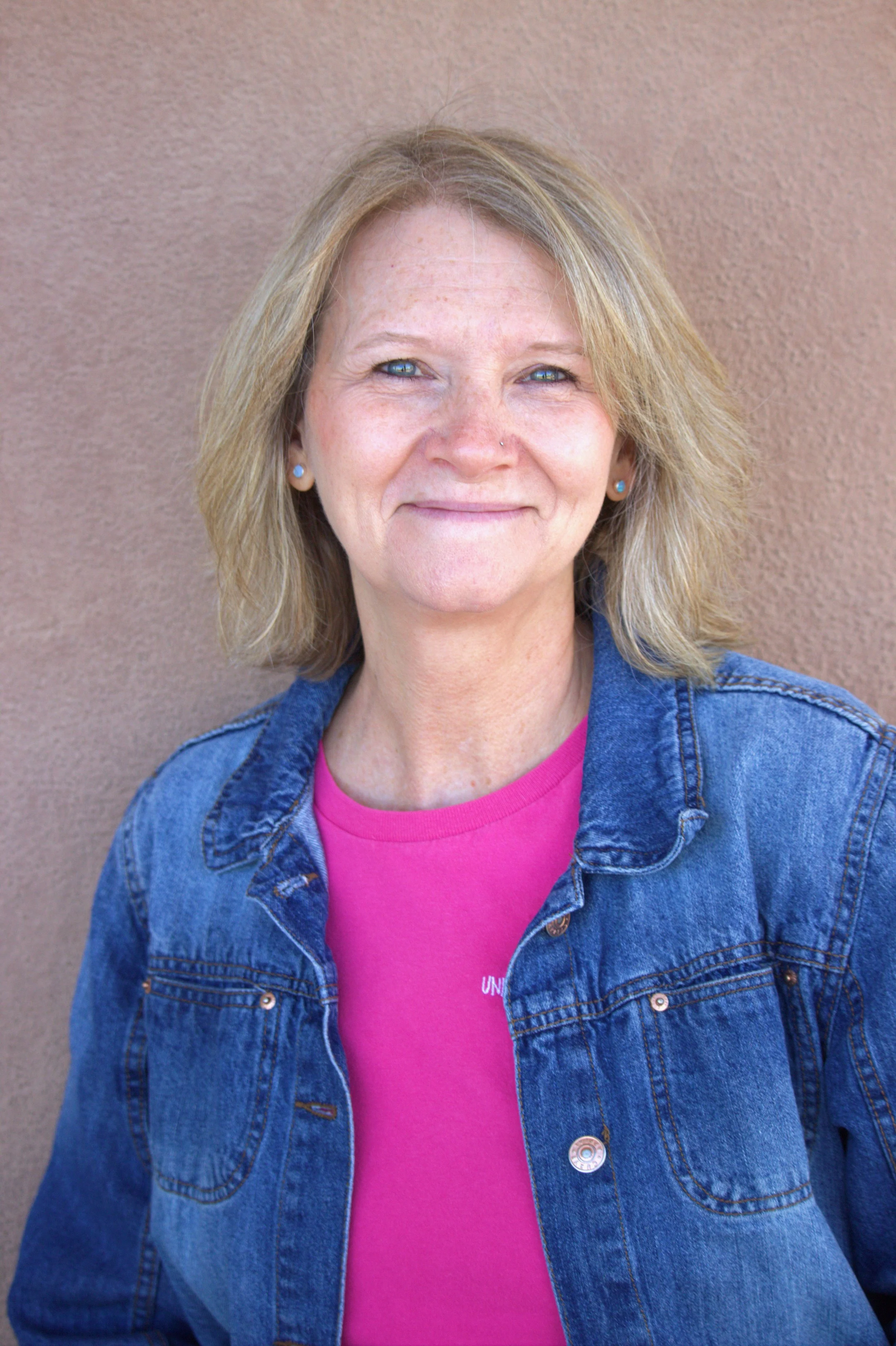 Portrait of a woman with blonde hair, blue eyes, and a subtle smile, standing against a brown textured wall.