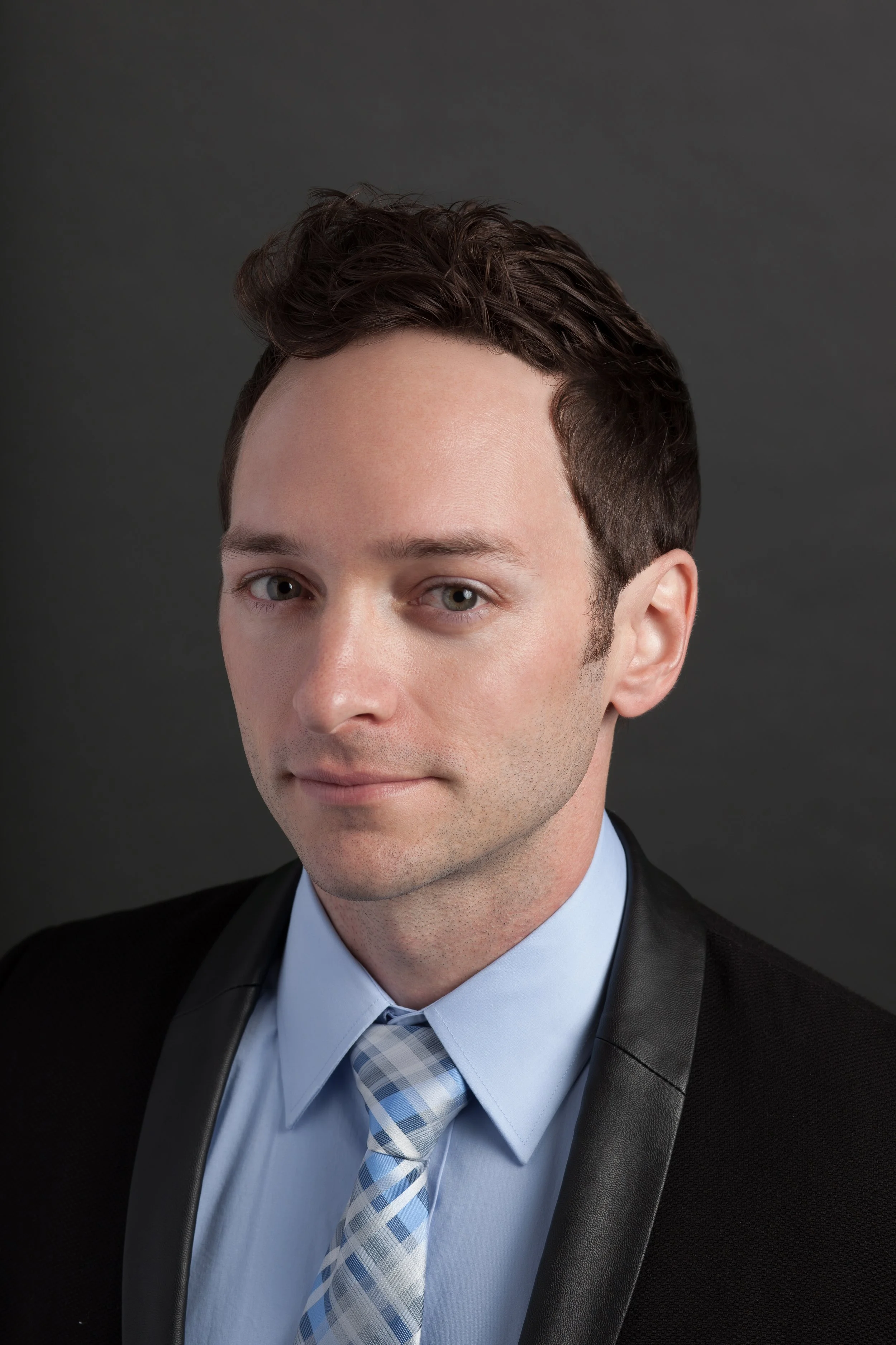 Portrait of a young man with short, dark hair wearing a light blue dress shirt, a patterned blue and white tie, and a black blazer, against a dark gray background.