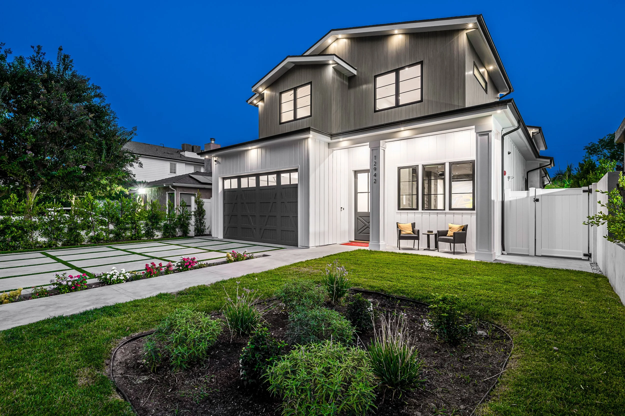 Modern two-story house with a white and gray exterior, front lawn, flower bed, and seating area outside during evening.