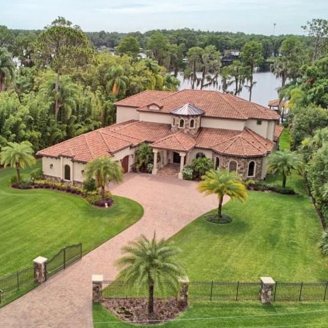 View of a large houses with red tile roof, surrounded by greenery and trees near a body of water.