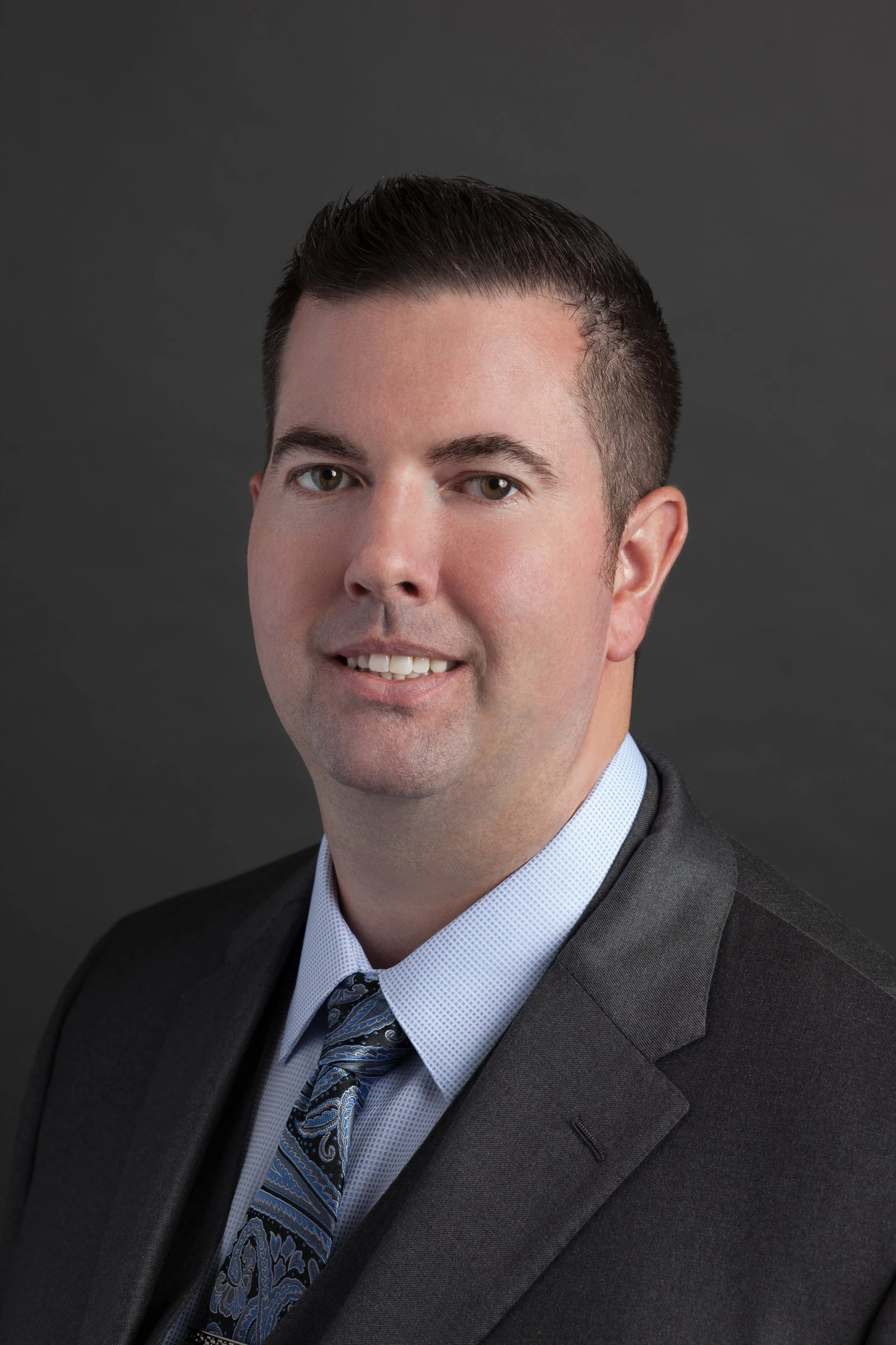 Professional headshot of a man wearing a dark suit, light dress shirt, and patterned tie, with a dark background.