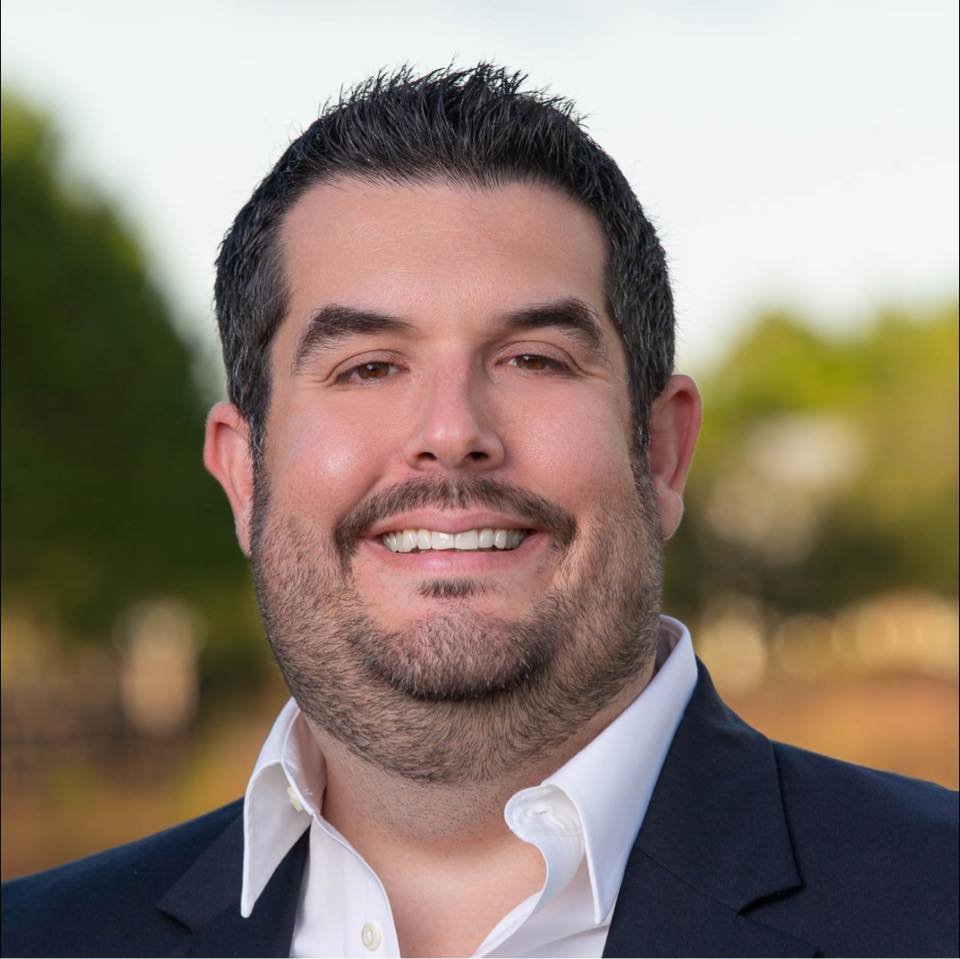 A smiling man with dark hair, facial hair, and a suit jacket, standing outdoors with a blurred background of trees and sky.