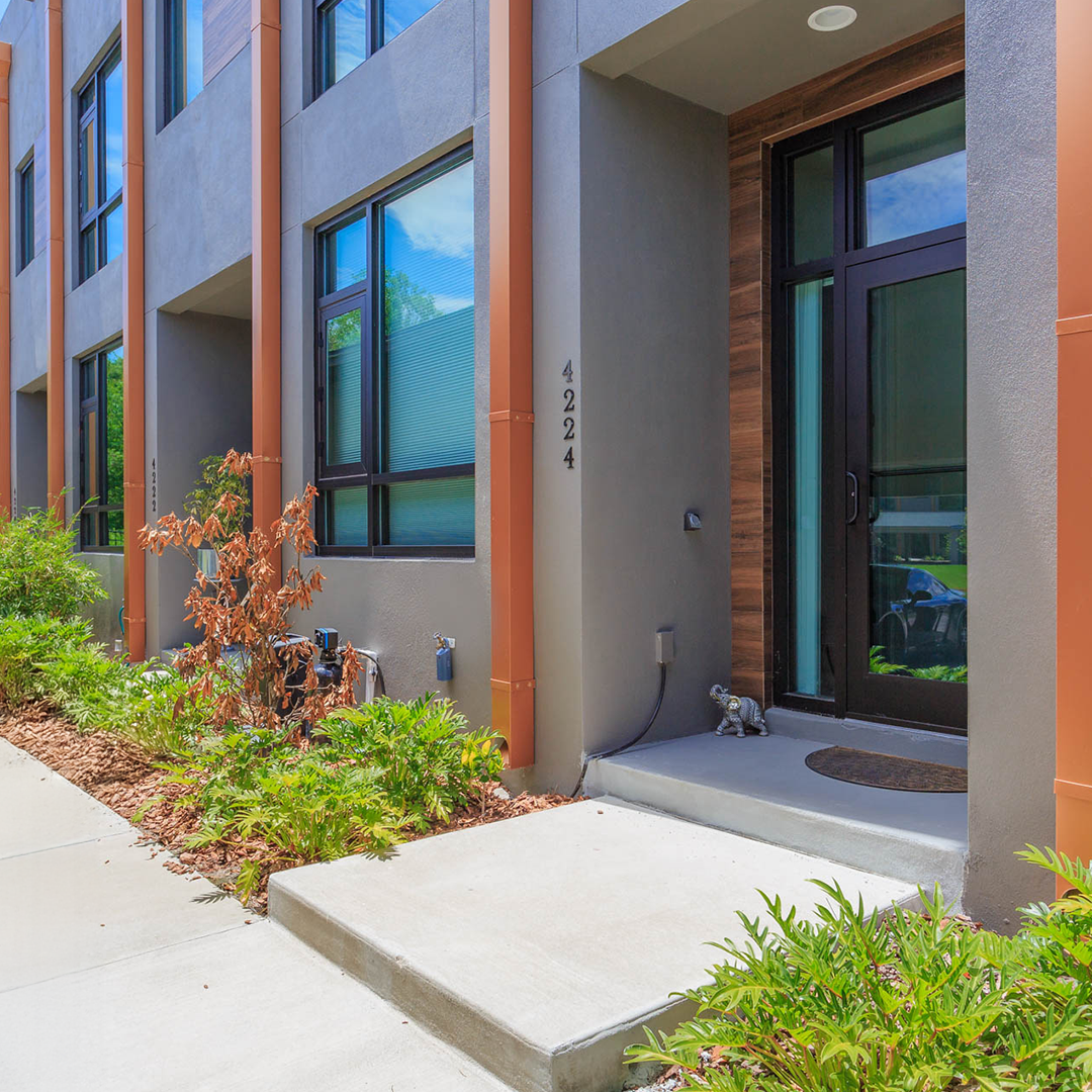 Modern apartment building entrance with gray walls, black window frames, and orange vertical accents. A small front porch has a doormat and a decorative tiger figurine. There is green shrubbery and plants along the sidewalk, and the building number "4224" is visible.