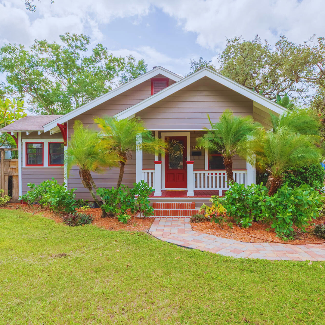 A cozy suburban house with a front porch, striped siding in gray and white, red accents on the door and window trim, surrounded by green palms, shrubs, and a lawn, with a brick pathway leading to the front steps.