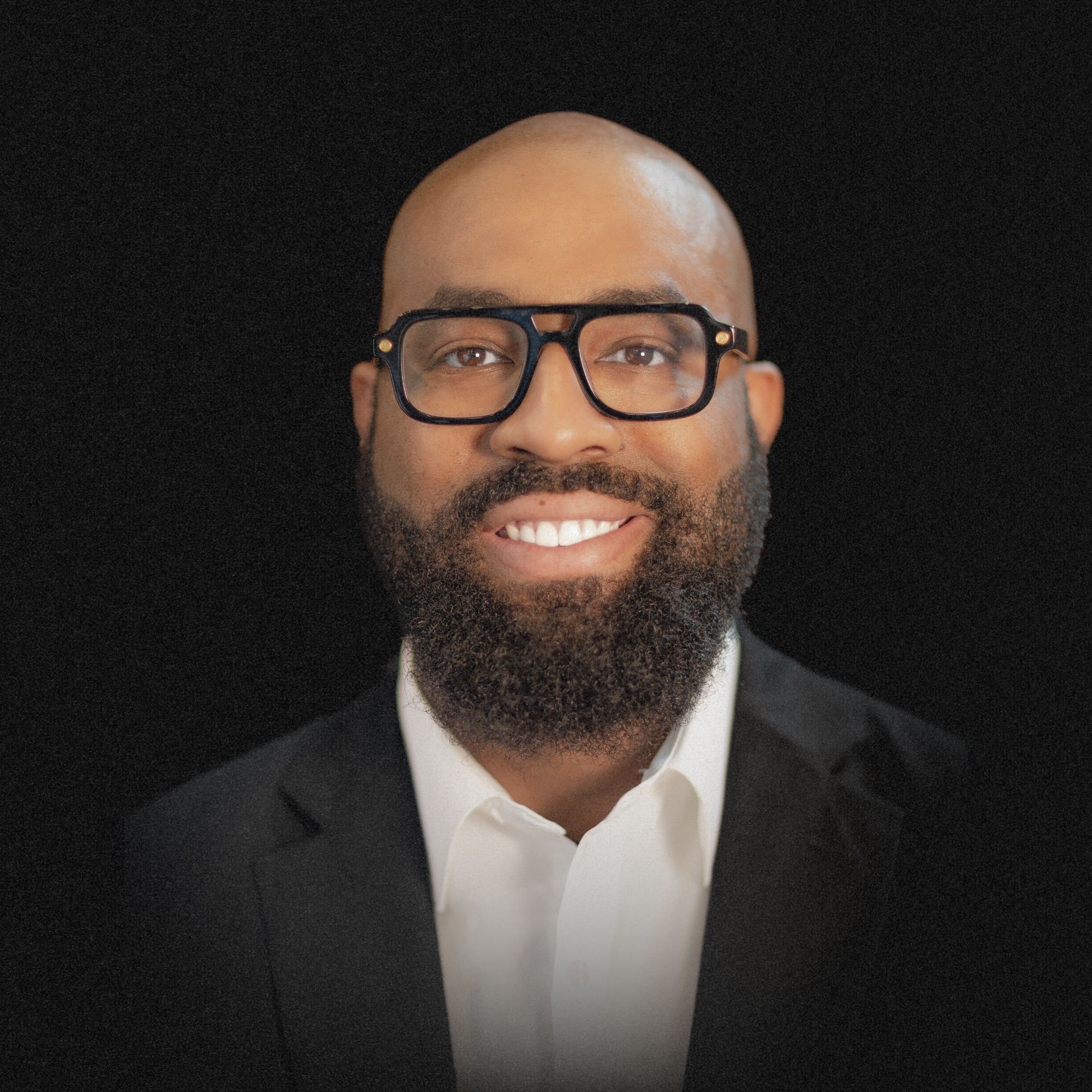 Headshot of a smiling man with glasses, a beard, wearing a black suit and white dress shirt, against a black background.
