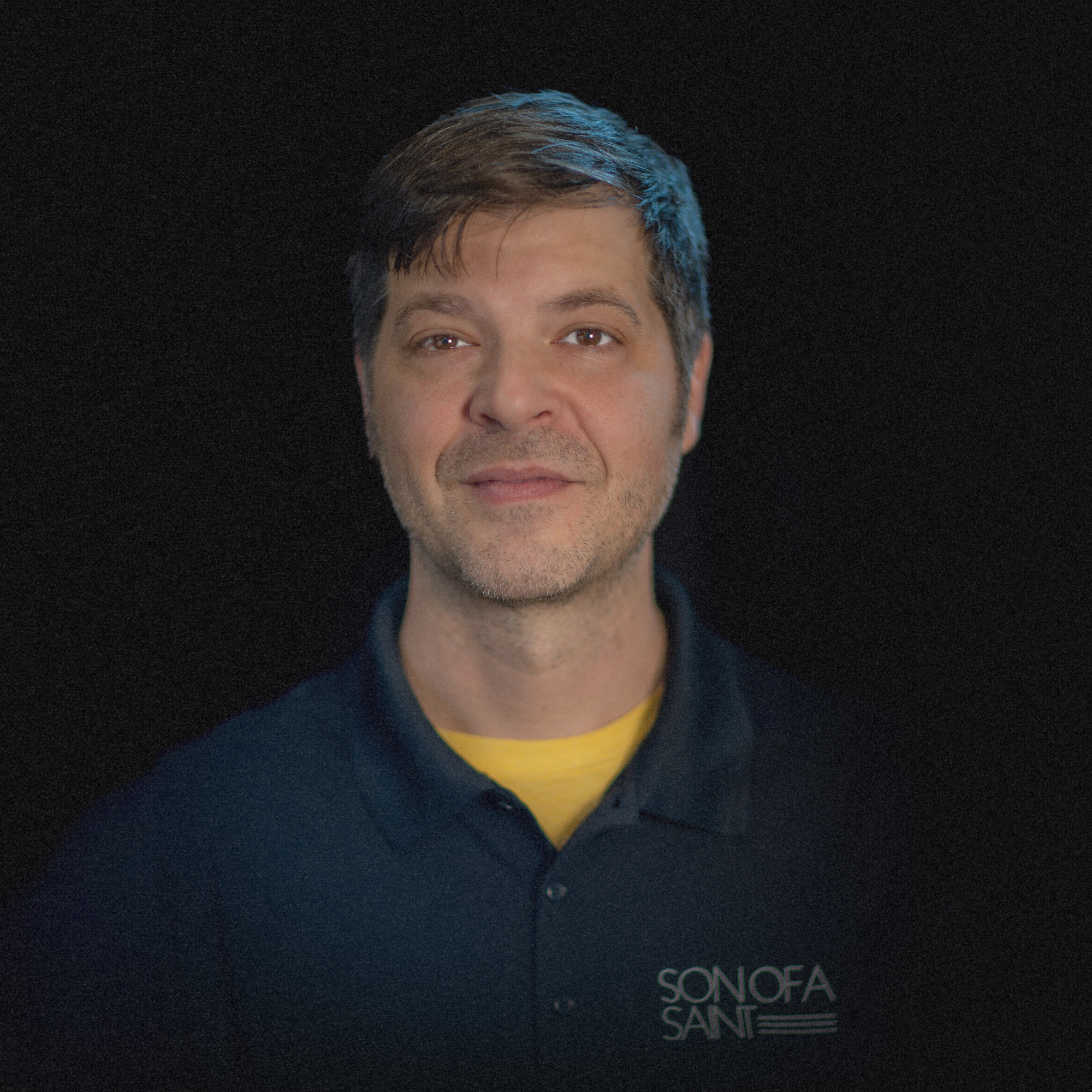 Portrait of a man with light skin, dark brown hair, and a light beard, wearing a black polo shirt with a yellow t-shirt underneath and the logo 'Sonofa Sandi' on it, against a black background.